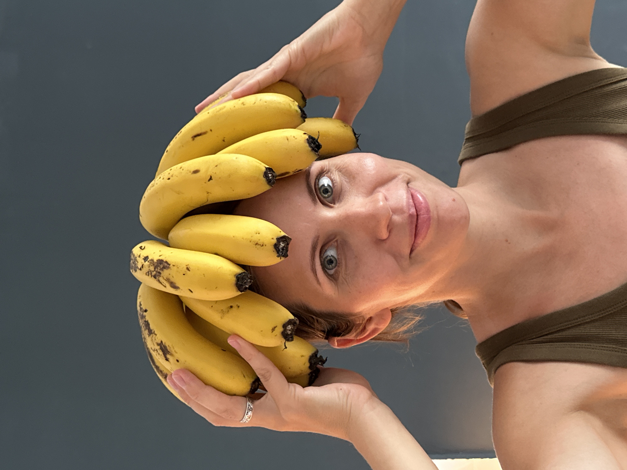 A woman is posing for a portrait with a bunch of bananas on her head. She is wearing a green tank top and has a silver ring on her left hand. The background is a plain dark wall. There is no indication of the image being taken in Fethiye, Turkey.