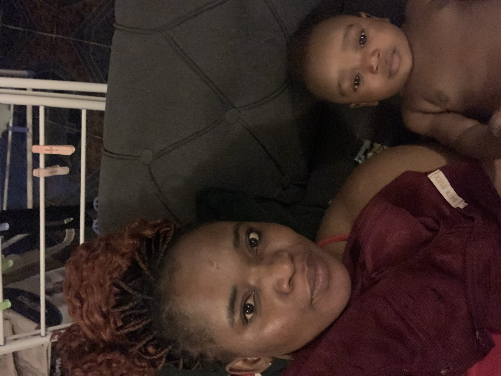 A woman with red braided hair and a burgundy shirt lies on a dark gray tufted cushion, near a baby with dark skin. The setting appears to be indoors. A white metal structure is visible to the left. There is no indication of the city or country.