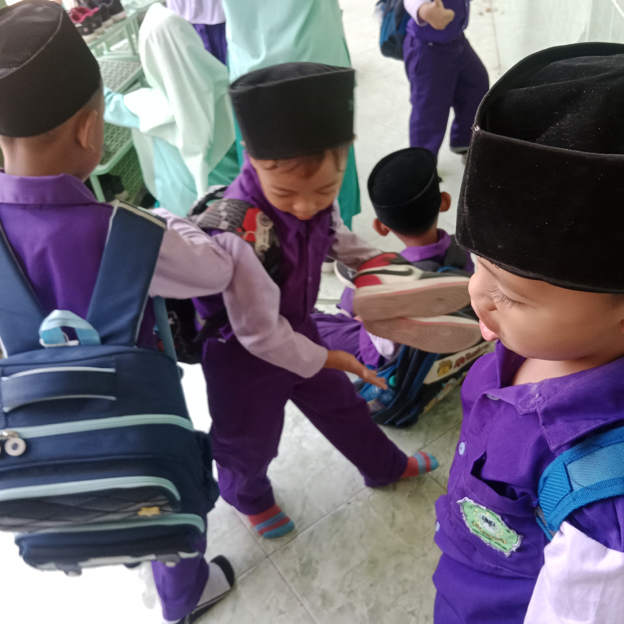 The image shows a group of young children, likely students, in what appears to be an outdoor school setting in Tegowanu Kulon, Indonesia. The children are wearing matching purple uniforms and black peci caps. One child in the foreground is facing away from the camera, wearing a dark blue backpack. Another child is visible from the back, also in a purple uniform and peci. A third child, facing towards the camera, is partially obscured by another child and appears to be reaching out. Their footwear includes striped socks and athletic shoes. The ground is tiled with a light-colored pattern. In the background, other children in similar attire and light-colored clothing are visible, along with some green and white structures that might be part of the school building or playground equipment. The overall scene suggests a casual school day activity.