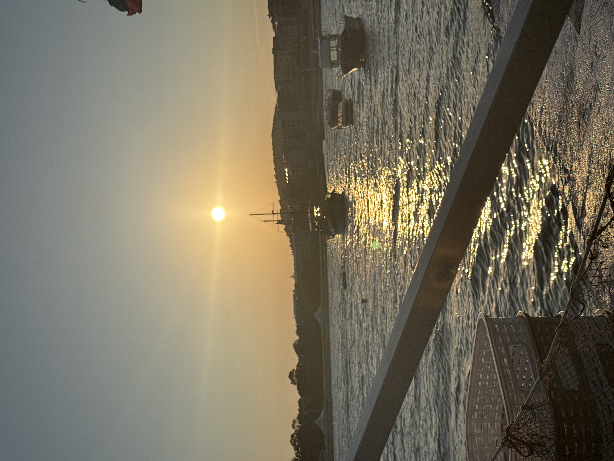 The image depicts a tranquil seascape at sunset, likely in Primorsko, Bulgaria, given the context. The sun, a bright disc, hangs low in the sky, casting a golden glow across the water and illuminating the scene with a warm, soft light. The sky transitions from a pale blue at the top to a warmer, orangish hue near the horizon, a typical characteristic of a sunset.
Several boats are visible on the water. To the left, a traditional sailing vessel with a tall mast stands out. Closer to the center and right, smaller motorboats are moored, bobbing gently on the waves. The water itself is moderately choppy, with the sunlight creating a shimmering, sparkling effect on its surface.
In the background, a treeline forms a dark silhouette against the sky, with hints of buildings and structures nestled among the trees, suggesting a coastal town or resort. The overall atmosphere is serene and peaceful, evoking a sense of calm and natural beauty.
In the foreground, a white plastic basket and fishing nets are visible, resting on what appears to be the deck of a boat. A metal railing or beam extends diagonally across the lower part of the frame, partially obscuring the view of the water and boats, indicating that the photograph was taken from another vessel or pier. The presence of the basket and nets suggests a fishing or maritime context.