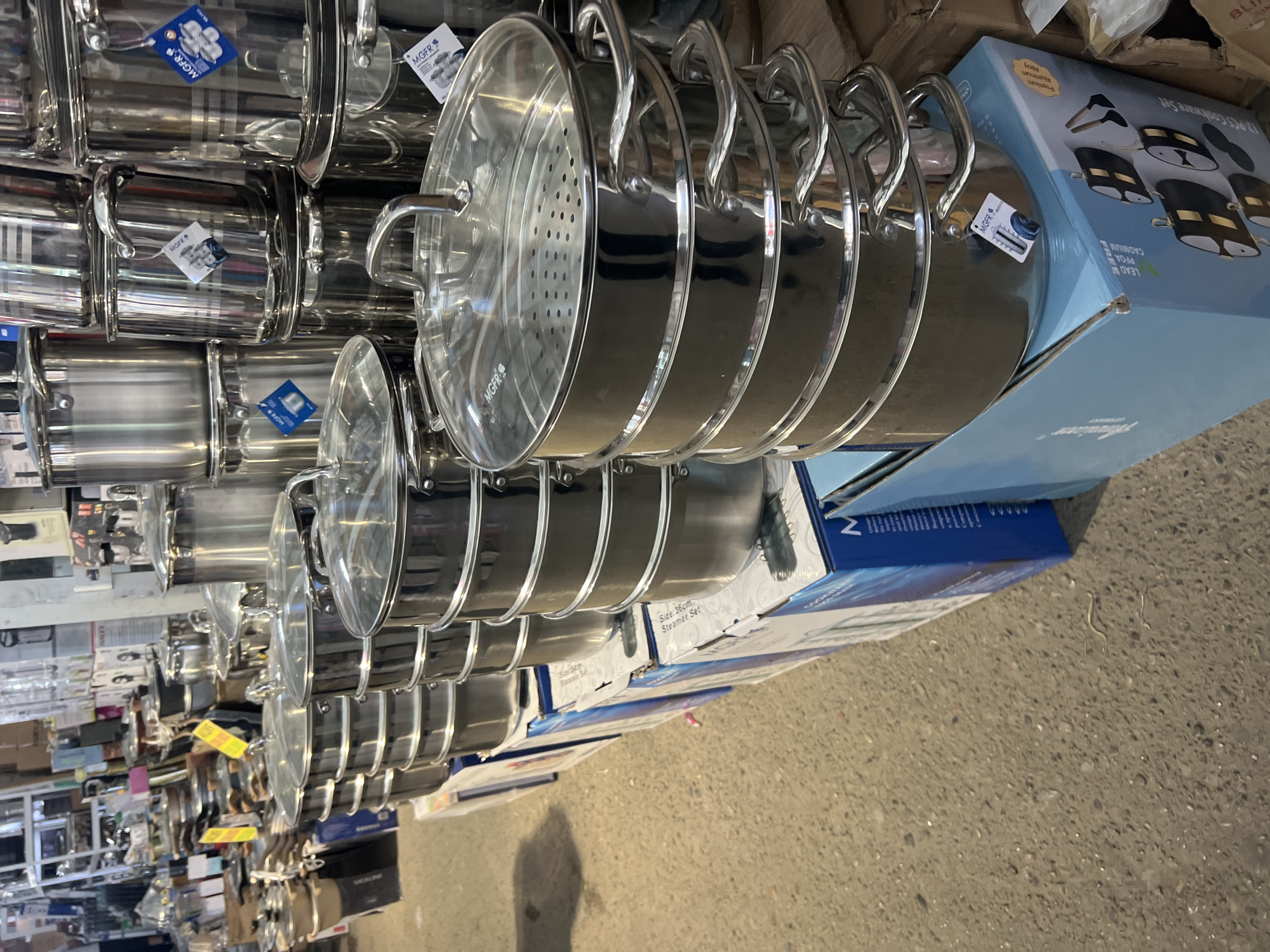 The image shows a retail display of stainless steel cookware, specifically steamers, in a store. The steamers are stacked high on top of blue boxes. The boxes have graphics and text indicating a 12-piece cookware set and features like being lead, PFOA, and cadmium-free. The store appears to be in a market or warehouse setting with a concrete floor and other merchandise visible in the background. The overall impression is that of a busy commercial environment. The city of Eshanguzar, Uzbekistan is mentioned as the location, but there are no visual cues in the image to confirm this specific location.