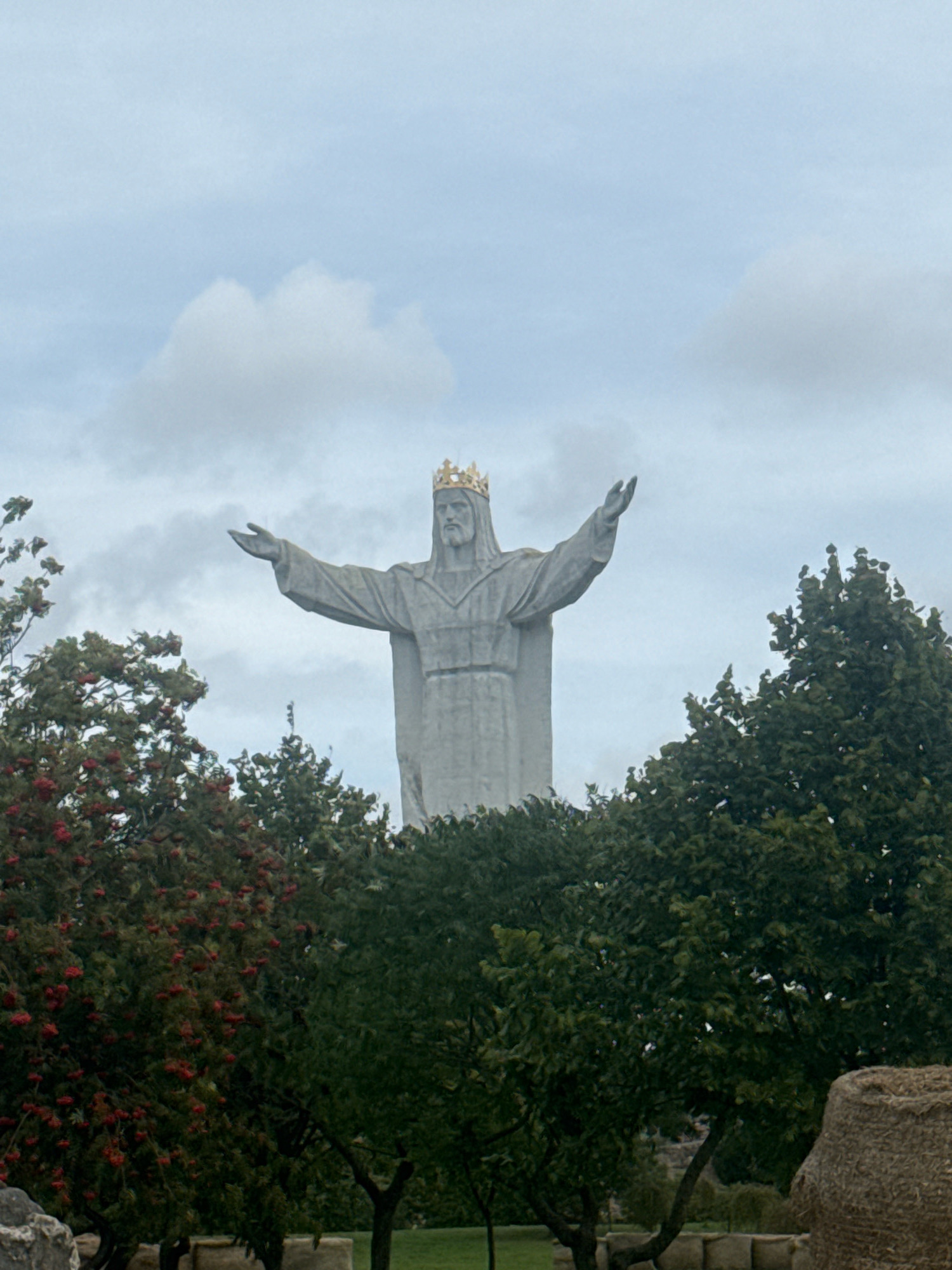 The image captures a large, gray, stone statue of Jesus Christ, known as Christ the King, located in Świebodzin, Poland. The statue is positioned centrally in the frame, elevated on a pedestal, and is distinguished by a golden crown atop its head. Christ's arms are outstretched, with the right arm extending slightly upwards and the left arm pointing outwards. The statue's face, though somewhat indistinct due to distance and image quality, appears to have a beard. The statue is partially obscured by lush green trees and shrubs in the foreground. Some of these trees bear clusters of red berries. The background consists of a cloudy, pale blue sky. The overall scene is one of a monumental religious statue set within a natural, outdoor environment.