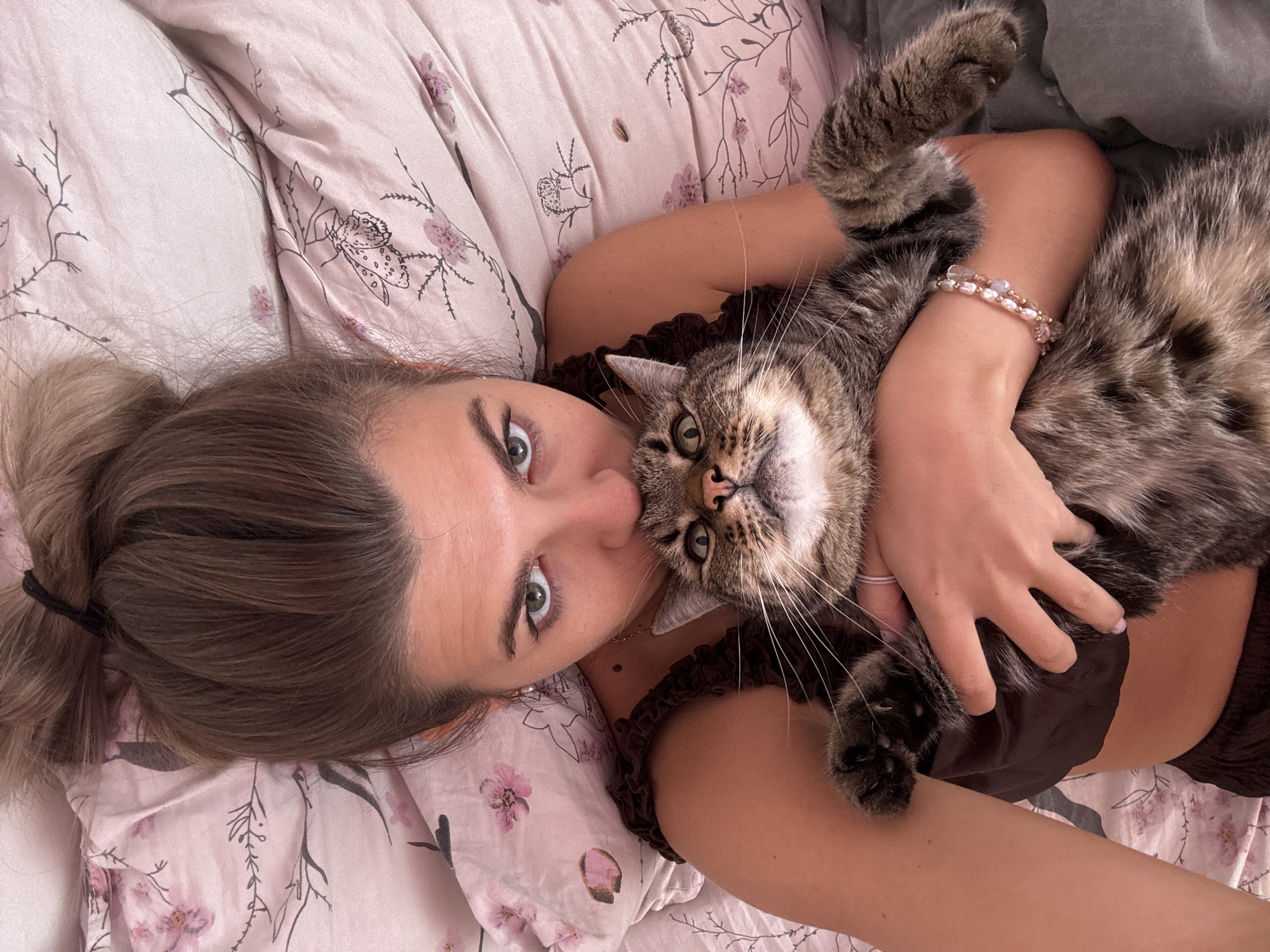 In a close-up shot, a young woman with her hair in a bun and wearing a brown camisole is lying in bed, holding a tabby cat. The woman is kissing the cat on the forehead. She has striking green eyes and appears to be looking directly at the camera. The bedsheets are pink with a floral pattern. The overall impression is one of affection and comfort. There is no discernible information to place this scene in Chaikovsky, Russia.