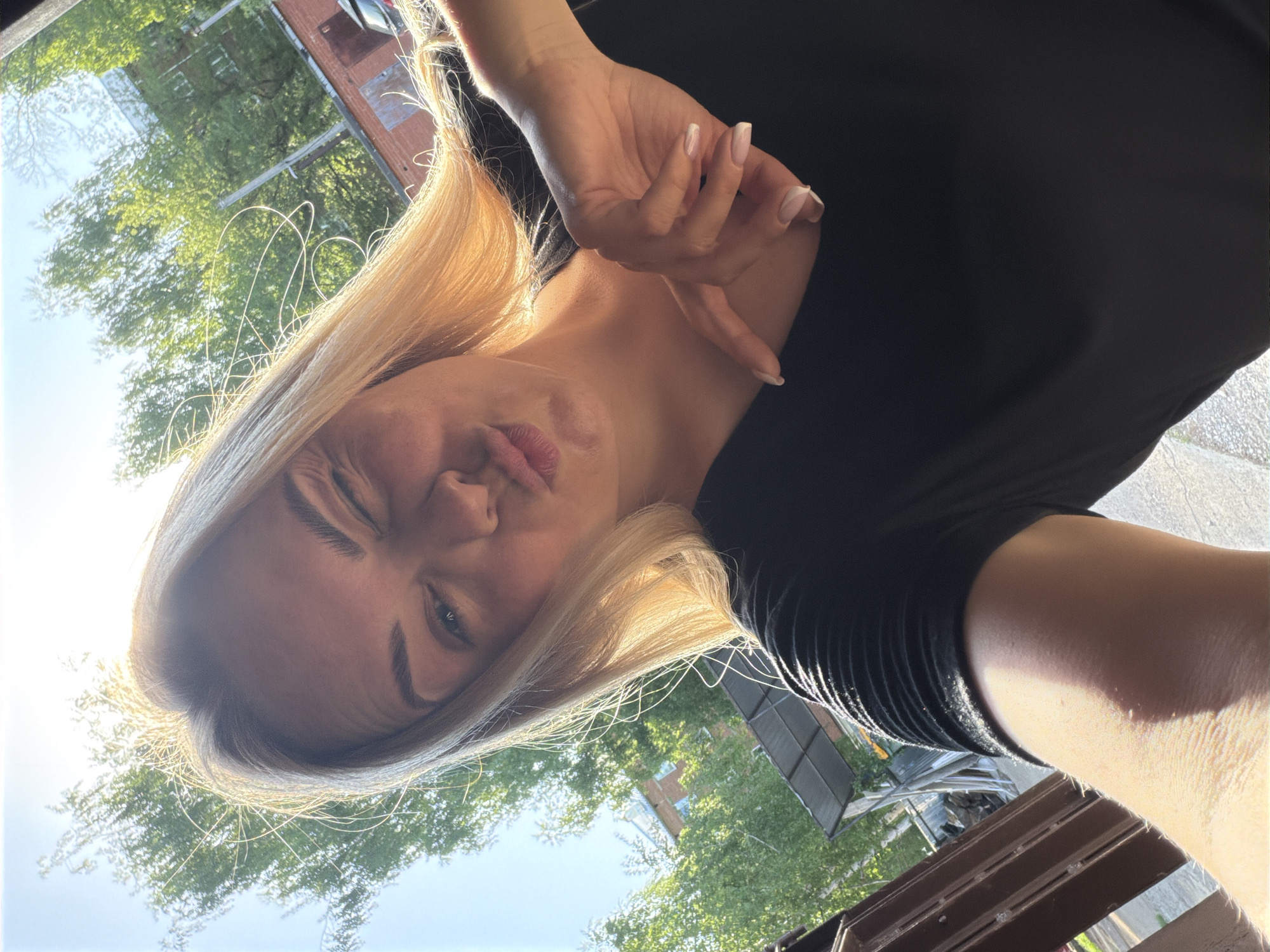 A woman with blonde hair is taking a selfie outdoors. She is wearing a black shirt. Her right eye is closed in a wink, and her lips are puckered in a kiss. Her left hand is raised near her chest with fingers slightly curled. In the background, there are trees and buildings, possibly indicating an urban area. Specific location in Votkinsk, Russia is not determinable from the image.