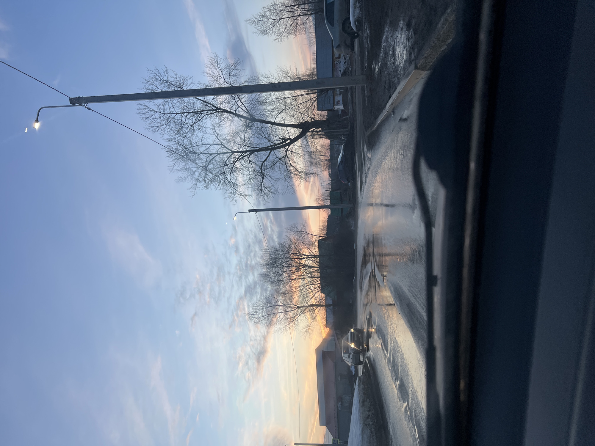 The image depicts a street scene in what appears to be the city of Chaikovsky, Russia, observed through a wet window, possibly a car window. The sky shows a sunset or sunrise with a mix of blue and orange hues. Bare tree branches are silhouetted against the sky. A street light illuminates the scene, and power lines stretch across the view. The road is wet, reflecting the sky and surrounding objects. A car is visible on the road. Buildings line the side of the street, suggesting an urban or suburban environment. The general atmosphere suggests a cold or damp climate.