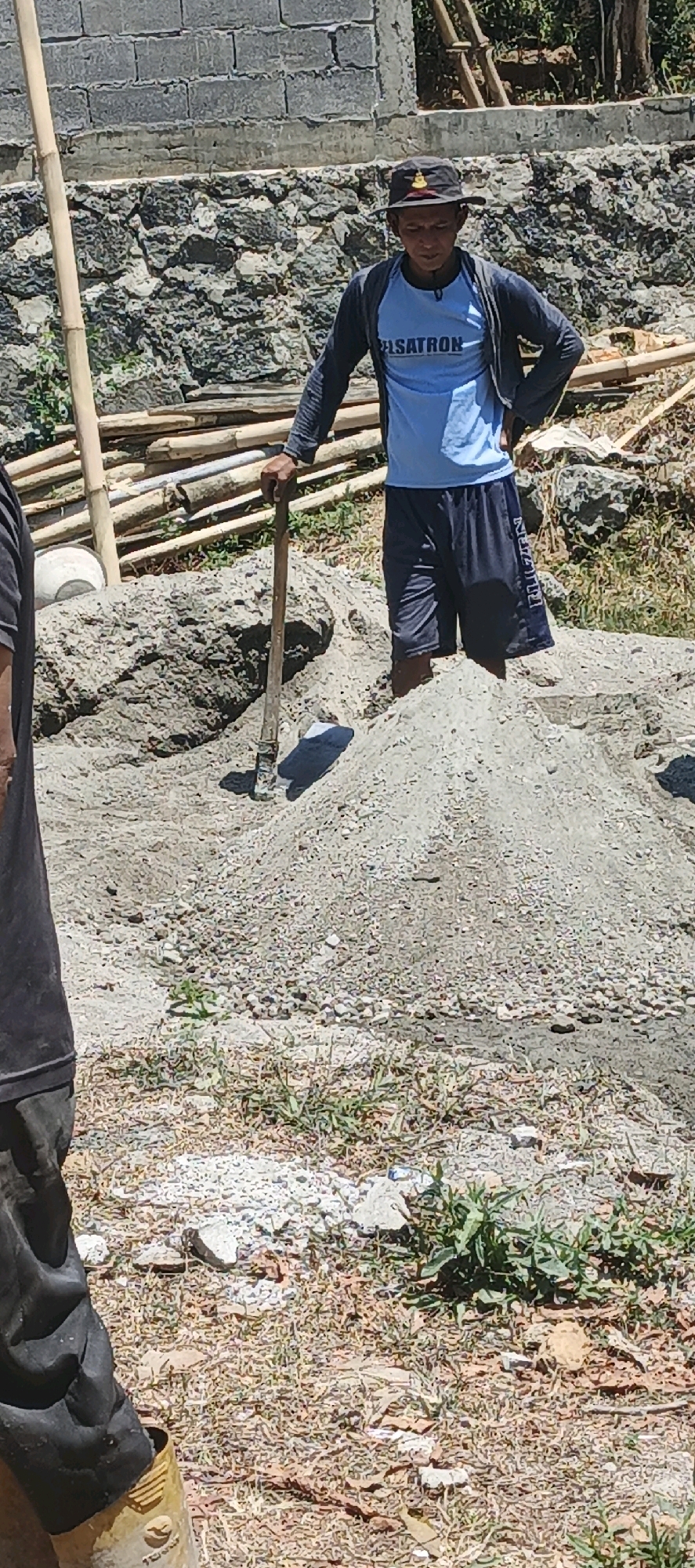 A man wearing a light blue t-shirt, a dark gray jacket, dark shorts, and a black hat stands in a pile of gravel. He is holding a shovel. He is in a construction area with piles of gravel and sand, bamboo poles, and a stone wall in the background. There is also a person on the left side of the image wearing a dark shirt and dark pants. The location is Jlarem, Indonesia.