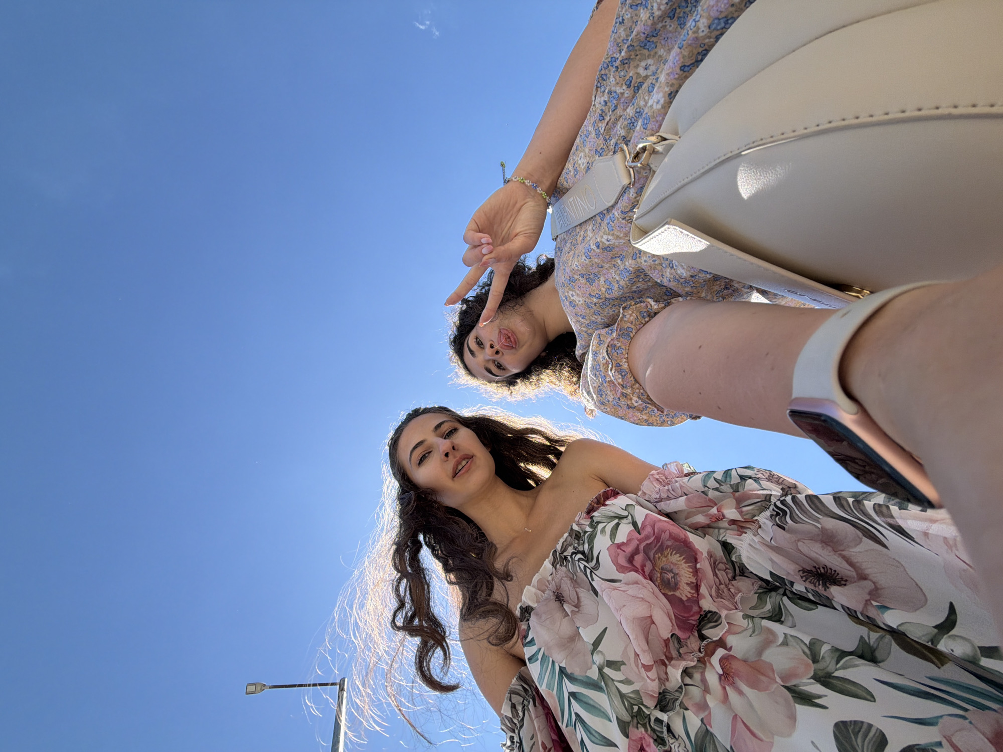 Two young women are captured from a low-angle perspective against a bright blue sky. The woman on the left, with long, dark, wavy hair, wears an off-the-shoulder floral dress with large pink and white blossoms. Her gaze is directed towards the camera. The woman on the right, also with dark curly hair, is sticking her tongue out and making a peace sign with her fingers. She is wearing a floral-patterned dress and carrying a light-colored handbag. A white smartwatch is visible on the arm of the person taking the photo. In the background, a street lamp and some wispy, dry grass are visible. The scene suggests a sunny day, possibly in an outdoor urban setting. While specific details about the city of Sofia, Bulgaria are not explicitly present in the visual information of the image itself, if this image were to be hypothetically located in Sofia, it could be a park or an open area within the city on a clear day.