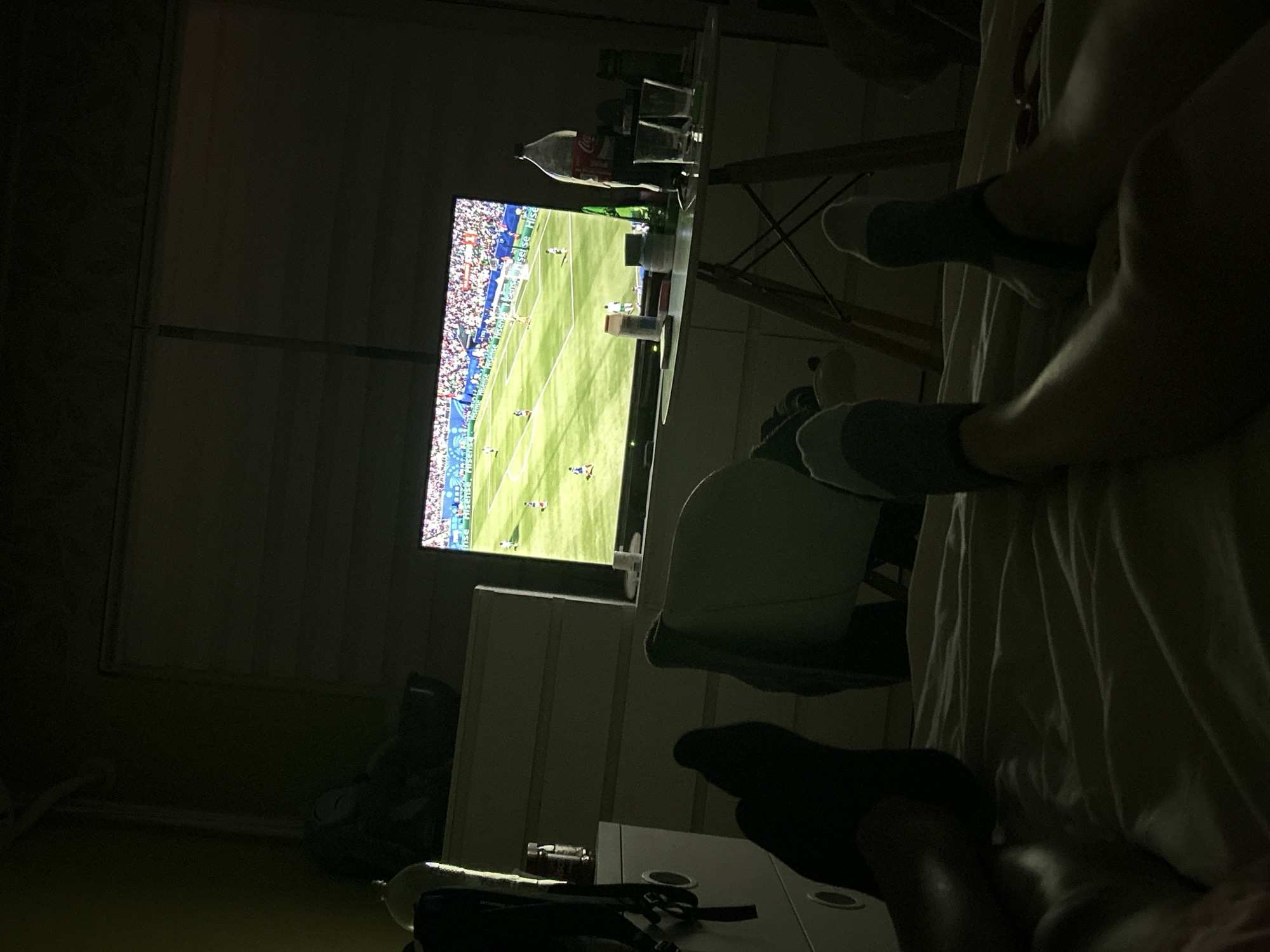 The image captures a low-angle, dimly lit scene of two individuals watching a soccer match on a television in a bedroom. The camera is positioned on a bed, focusing on the feet and lower legs of the individuals, who are wearing socks. The television screen, displaying the soccer game, is visible in the background, positioned on a table. Other objects present include a large bottle of Coca-Cola, glasses, and various containers on the table. To the left, a dresser with drawers and a backpack are visible. The overall environment suggests a relaxed setting in a private residence. The provided information about the location being Sofia, Bulgaria, is not visually verifiable from the image content.