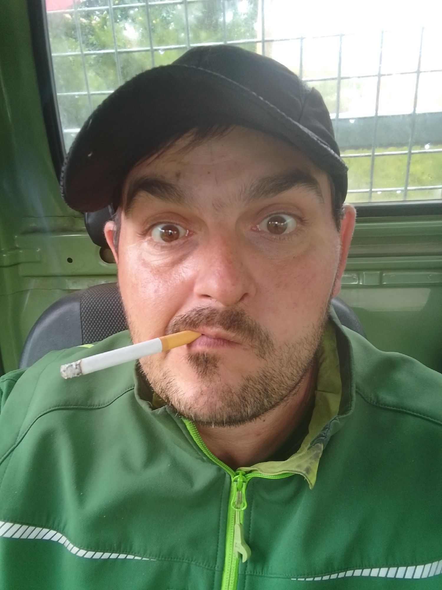 A man with brown eyes, a goatee, and a black cap is pictured from the chest up, inside what appears to be a vehicle cab. He is wearing a green jacket with a lime green zipper. A cigarette is held between his lips. The background consists of a window with a grid pattern, through which greenery is visible, suggesting an outdoor environment. The context of Hamburg, Germany is not discernible from the image itself.