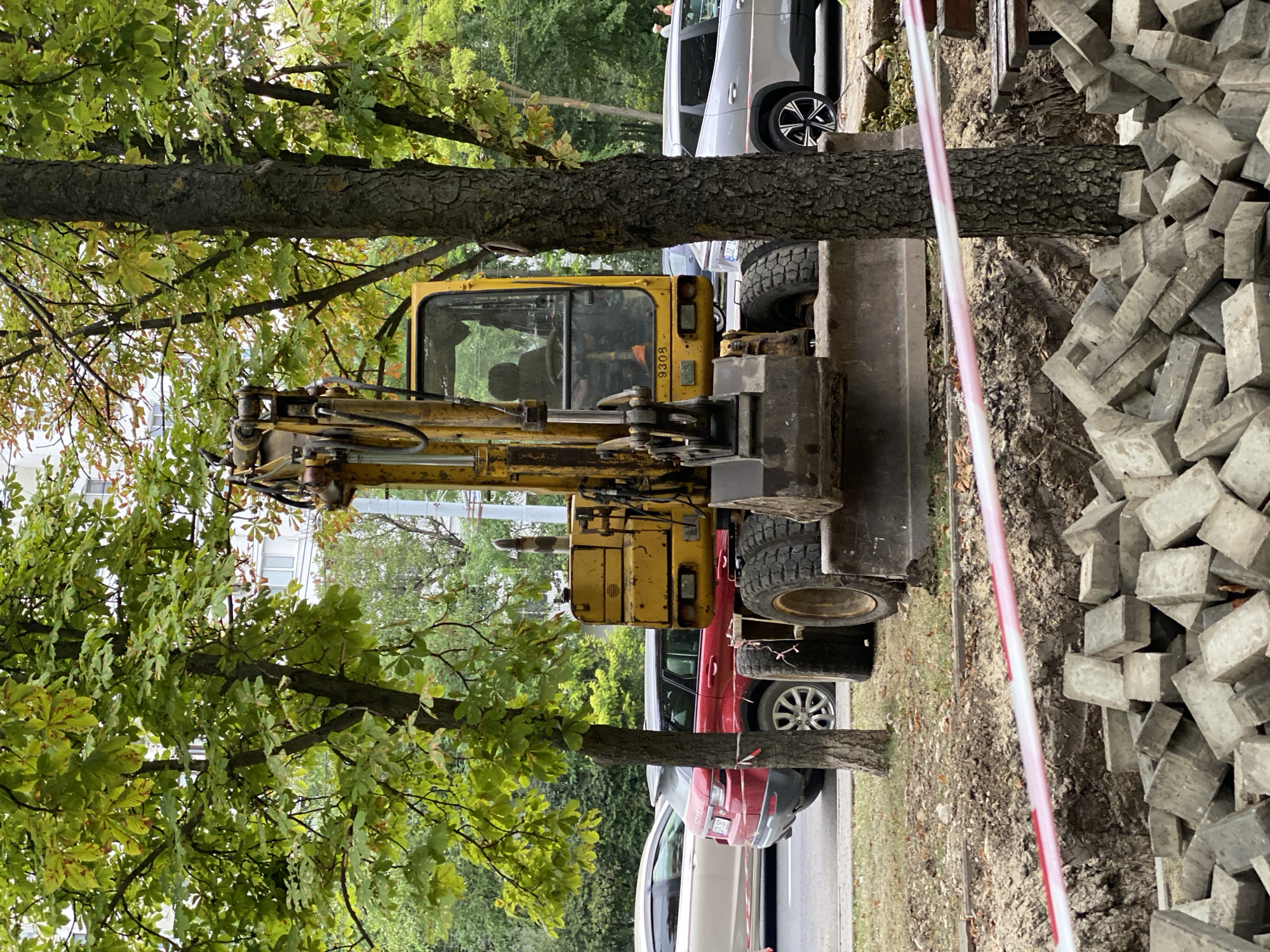 An excavator is parked on a city street, partially obscured by trees. The excavator is yellow and appears to be undergoing or involved in construction work, as evidenced by a pile of paving stones in the foreground and a trench with caution tape. Several cars are parked on the street behind the excavator, including a red SUV and a silver sedan. The background shows a building and more trees, indicating an urban environment. The presence of construction materials suggests ongoing road or sidewalk work. The general scene depicts a typical urban street scene with construction activity.