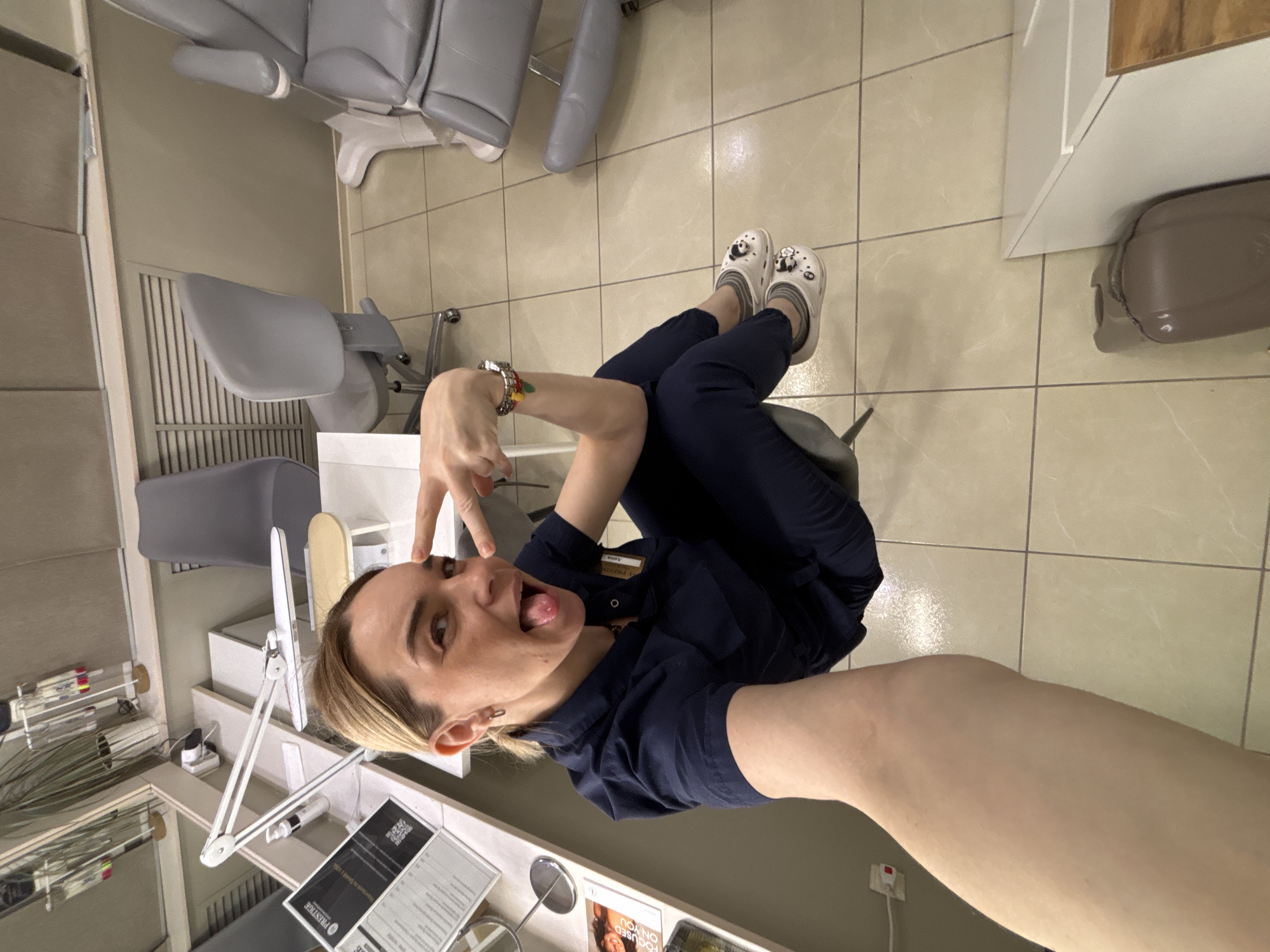 Here is a detailed description of the image:
A person with fair skin and short blonde hair is sitting in a chair in a room in Unknown, Russia. The individual is wearing a navy blue uniform and white slip-on shoes with cartoon panda decorations. They are sticking their tongue out and making a "peace" sign with their fingers near their face, holding the camera at an angle for a selfie.
The room appears to be a beauty salon or medical clinic. There is a grey dental chair, a computer monitor, and various pieces of equipment on a counter, including tweezers and small jars. A mirror is also visible, reflecting some of the equipment. The floor is covered in light beige tiles. A brown object is visible on a shelf.