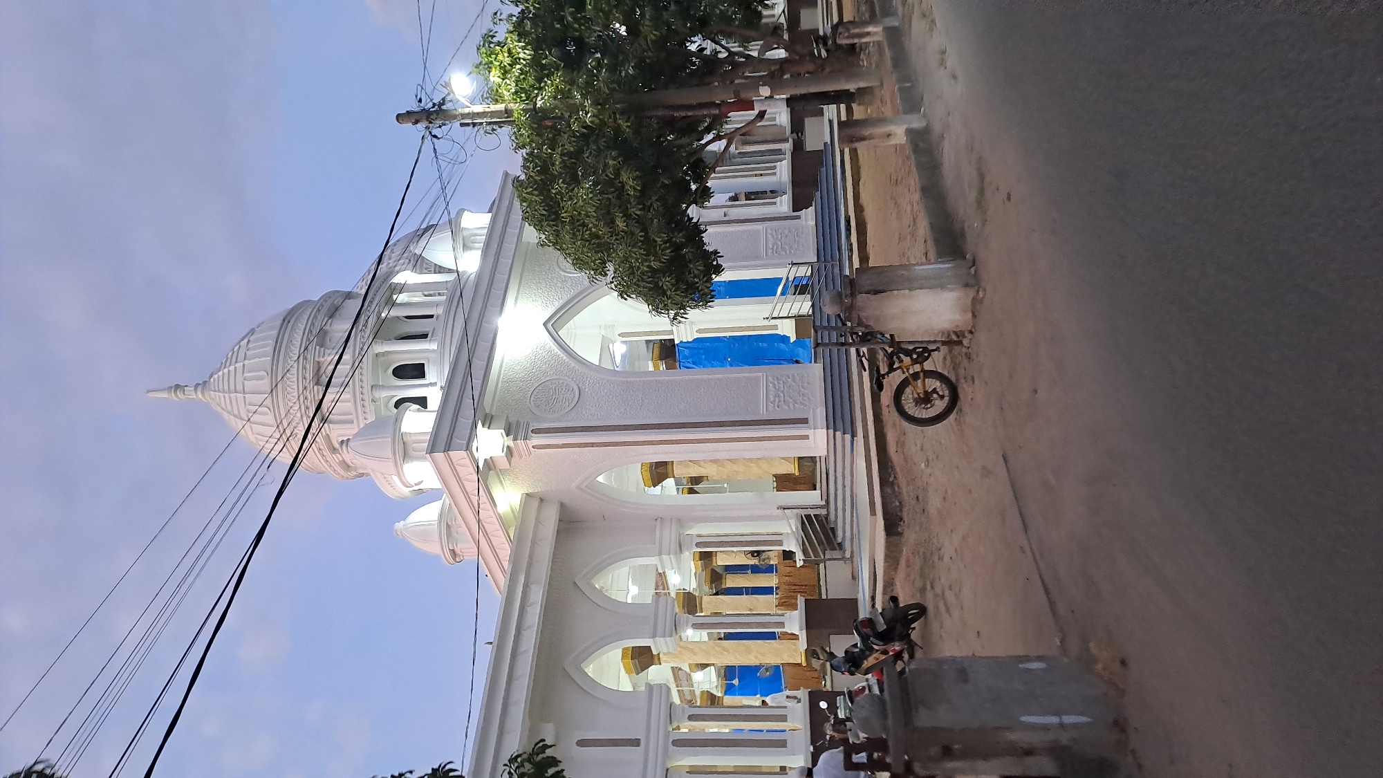 The image depicts a white building with multiple domes and arches, likely a mosque, located in the city of Lhokawe Teungoh, Indonesia. The building features intricate architectural details, including decorative carvings and columns. In front of the building, there is a paved area with several parked motorcycles and a bicycle. A large tree stands to the right of the building, and utility poles with overhead wires are visible in the background. The sky is partly cloudy, indicating it is either early morning or late evening. The overall scene suggests a public space within a community, possibly a religious or cultural center.