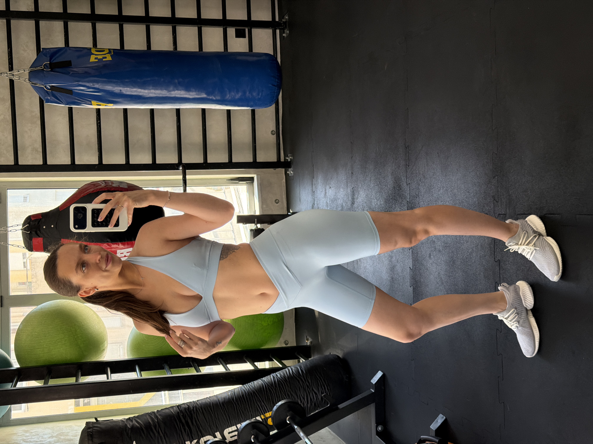 A woman in a light blue sports bra and matching biker shorts stands in a gym, holding a smartphone and taking a selfie. She has her hair in a ponytail and is wearing grey athletic shoes. The gym is equipped with various fitness equipment, including punching bags, medicine balls, and weight racks. The flooring is black rubber mats, and large windows offer a view of the city. The overall scene suggests a workout session or preparation for one.
