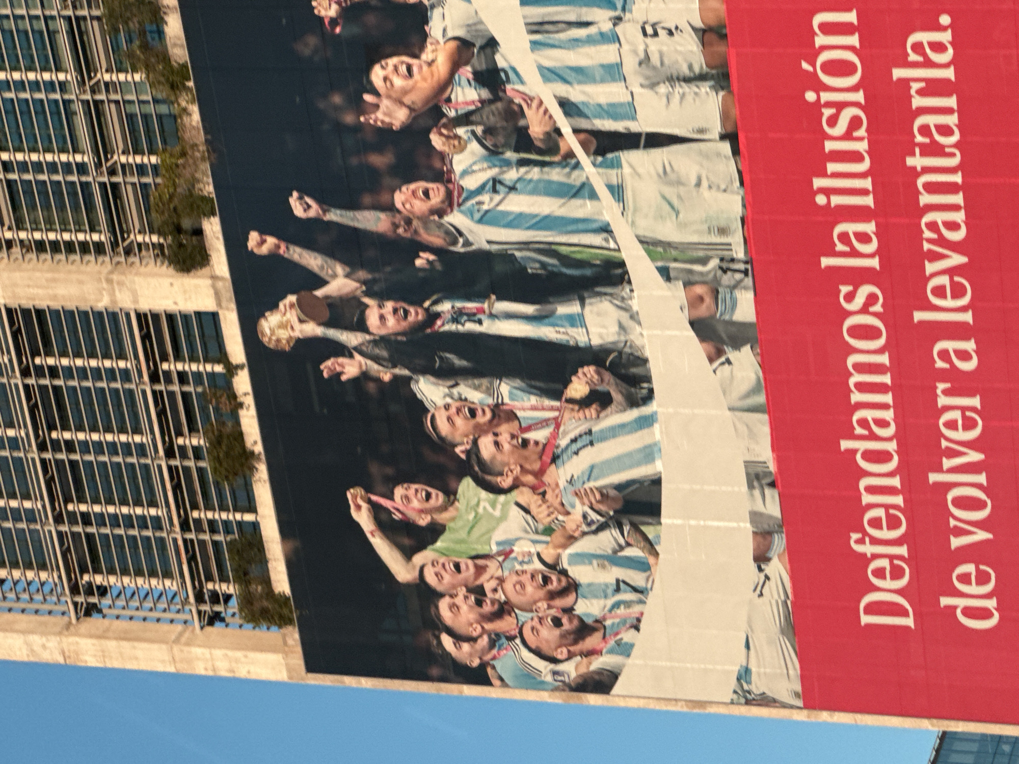 The photograph shows a large billboard attached to the side of a building in Buenos Aires, Argentina. The billboard features an image of the Argentinian national soccer team celebrating, wearing their white and blue striped jerseys and holding gold medals. The players are shown with their mouths open, likely shouting in celebration. To the right of the image, on a red background, is white Spanish text that reads, "Defendamos la ilusión de volver a levantarla." The building to which the billboard is attached has a modern facade with numerous windows and some greenery. The sky is visible in the lower left corner of the image, indicating a clear day.