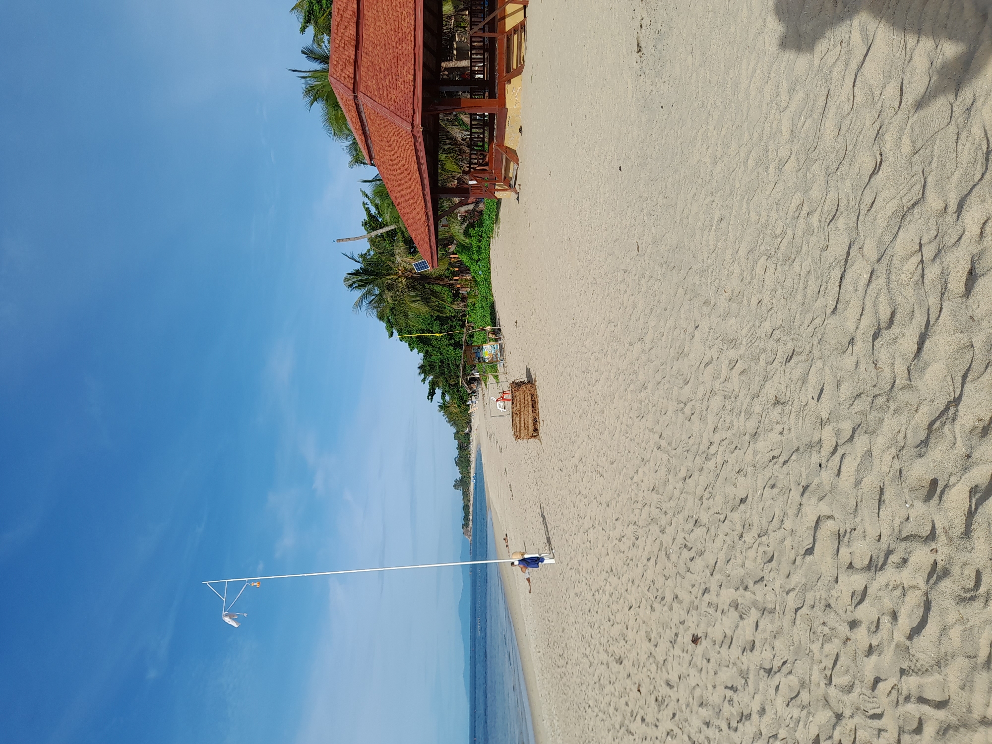 An eye-level, wide shot captures a tranquil beach scene. The sand, a light tan, is imprinted with numerous footprints and ripples, indicating the tide's recent presence. In the mid-ground, a white flagpole stands tall, adorned with a white flag and a small orange ornament. To the right of the flagpole, a person in a blue shirt and dark shorts walks away from the camera. Further to the right, a rustic, straw-covered structure sits on the sand, beside it, some people are reclining on chairs. Dominating the right side of the frame is a traditional wooden pavilion with a red, multi-tiered roof, supported by dark wooden posts, and accessed by two sets of wooden stairs. The pavilion is partially shaded by palm trees. In the background, the ocean stretches out, a calm expanse of blue meeting a distant, hazy horizon. The sky above is a vibrant, clear blue, with wispy white clouds. The scene is peaceful and serene, evoking a sense of vacation or relaxation.
