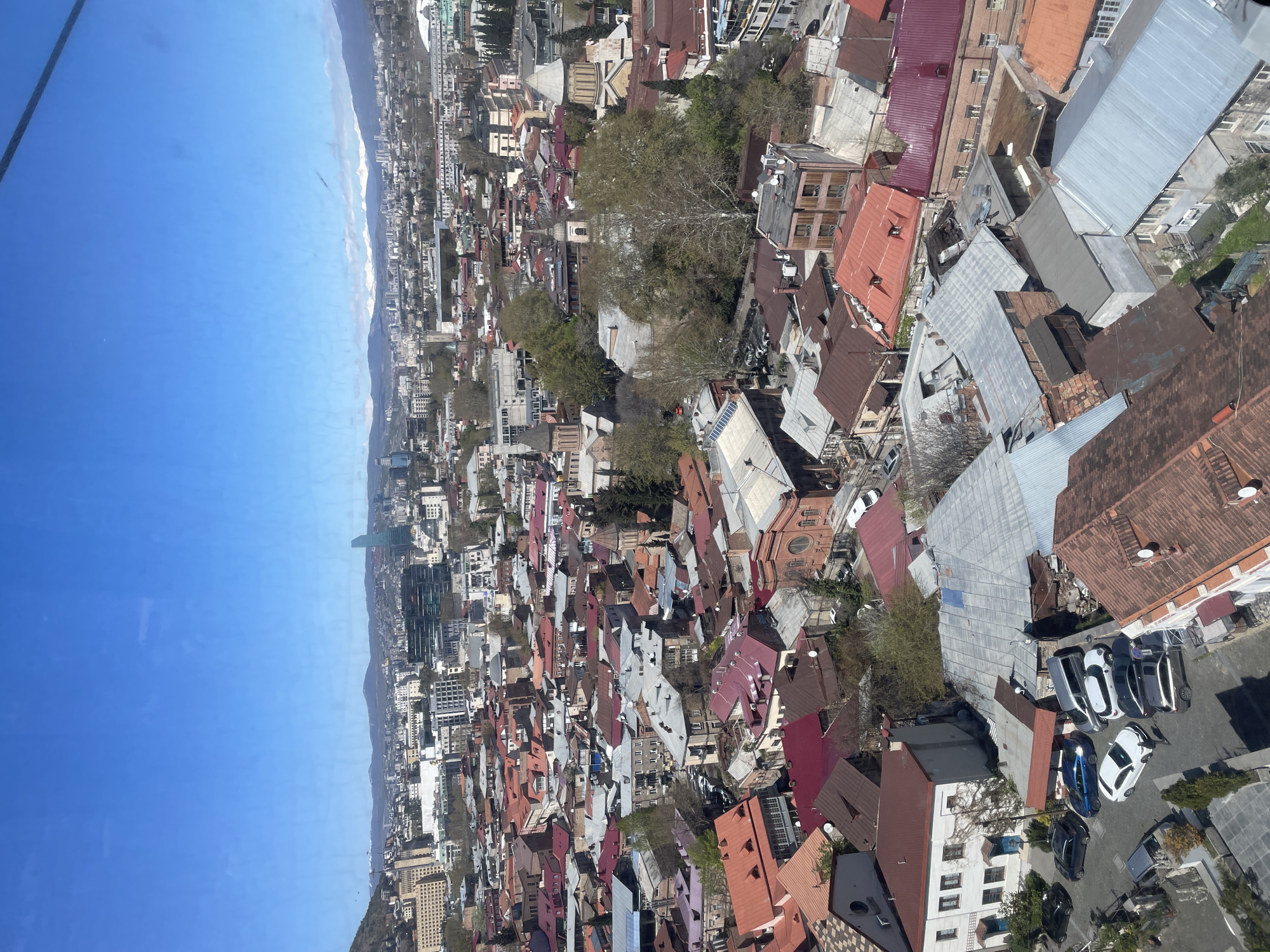 The image is an aerial view of Tbilisi, Georgia, looking towards the Black Sea in the distance. The scene shows a dense urban environment characterized by a mix of architectural styles and building heights, with a dominance of red-tiled roofs. To the left, the horizon reveals a sliver of the Black Sea under a clear blue sky. The city stretches out from the coast and is filled with a variety of buildings, from low-rise residential homes to mid-rise apartments. There are also some areas of trees, indicating some green spaces within the city. The roofs show the density of the city, and the overall impression is one of a vibrant, historical urban center. In the foreground, there are several cars parked in a courtyard, providing a perspective on the scale and the layout of the city from above. The angle of the picture suggests that it was taken from a high vantage point.