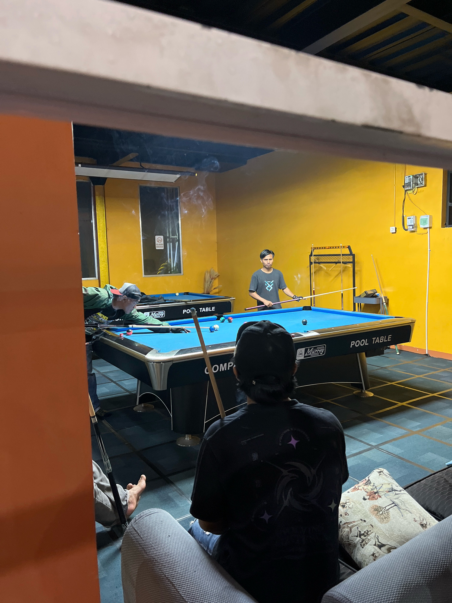 The image shows an indoor pool hall in Jatilawang, Indonesia, with a brightly lit yellow wall as the backdrop. In the foreground, the back of a person wearing a black t-shirt and a black baseball cap is visible, seated on a couch or chair, looking towards a pool table. The t-shirt has a dark, swirling graphic design with purple stars.
In the middle ground, a person wearing a green jacket and a baseball cap is bent over a blue pool table, holding a pool cue and in the process of playing. Another person, a young man in a grey t-shirt with a graphic logo, stands behind the same pool table, holding a cue and appearing to be either waiting for their turn or observing the game. The pool table is green and has several balls on it. There is a second pool table visible further back, also with a blue surface.
The environment appears to be a recreational space, possibly a bar or club, with lighting fixtures and electrical outlets visible on the yellow wall. There are also cues racks and a broom leaning against the wall in the background. Wisps of smoke are visible in the air, suggesting that smoking is permitted in this area. The floor is dark and tiled with a grid pattern. The image is taken from an angle that suggests the photographer is looking through an opening or doorway into the pool hall.