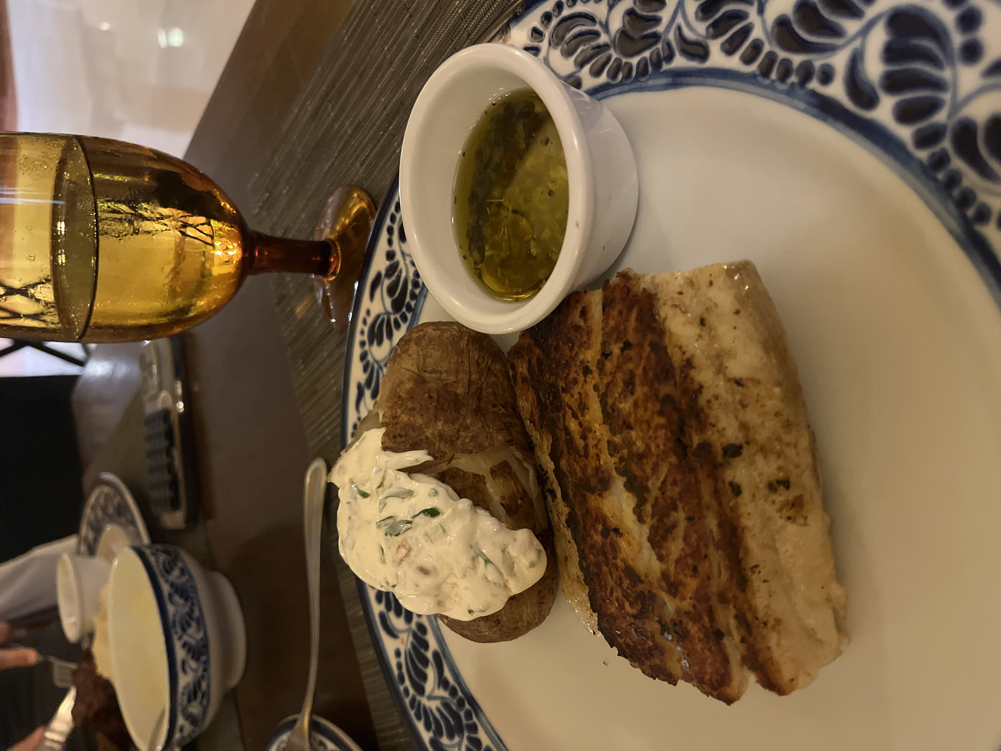 The image captures a close-up shot of a meal presented on a white plate with a distinctive blue floral pattern around the rim. The plate contains a baked potato topped with a creamy, herb-flecked sauce, a piece of grilled fish, and a small white bowl filled with a green, oily liquid, likely a dipping sauce or dressing. The meal is positioned next to a yellow-tinted glass with a brown stem, possibly containing a beverage. In the background, a portion of another plate or dish with similar blue patterns is visible, along with a silver fork. The table setting suggests an indoor dining environment, possibly a restaurant or private residence. It is likely that this image was taken in Playa del Carmen, Mexico.
