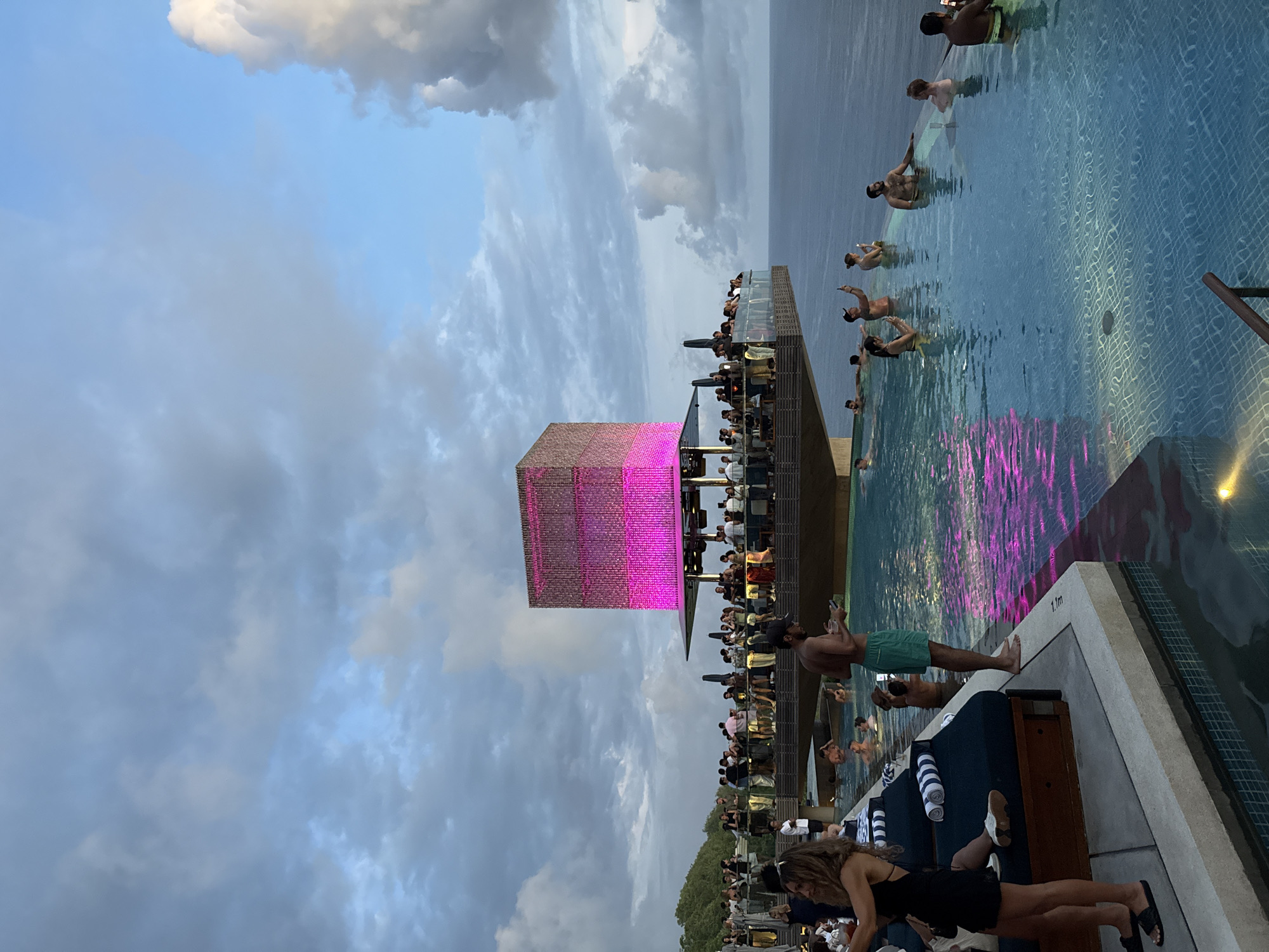The image shows a crowded outdoor infinity pool scene in Kuta Selatan, Indonesia, at sunset. The pool overlooks the ocean. People are swimming and standing in the pool and lounging on chairs around the deck. A large, illuminated pink cube structure stands on the edge of the pool deck. There are buildings and foliage in the lower section of the image. The sky is partly cloudy.