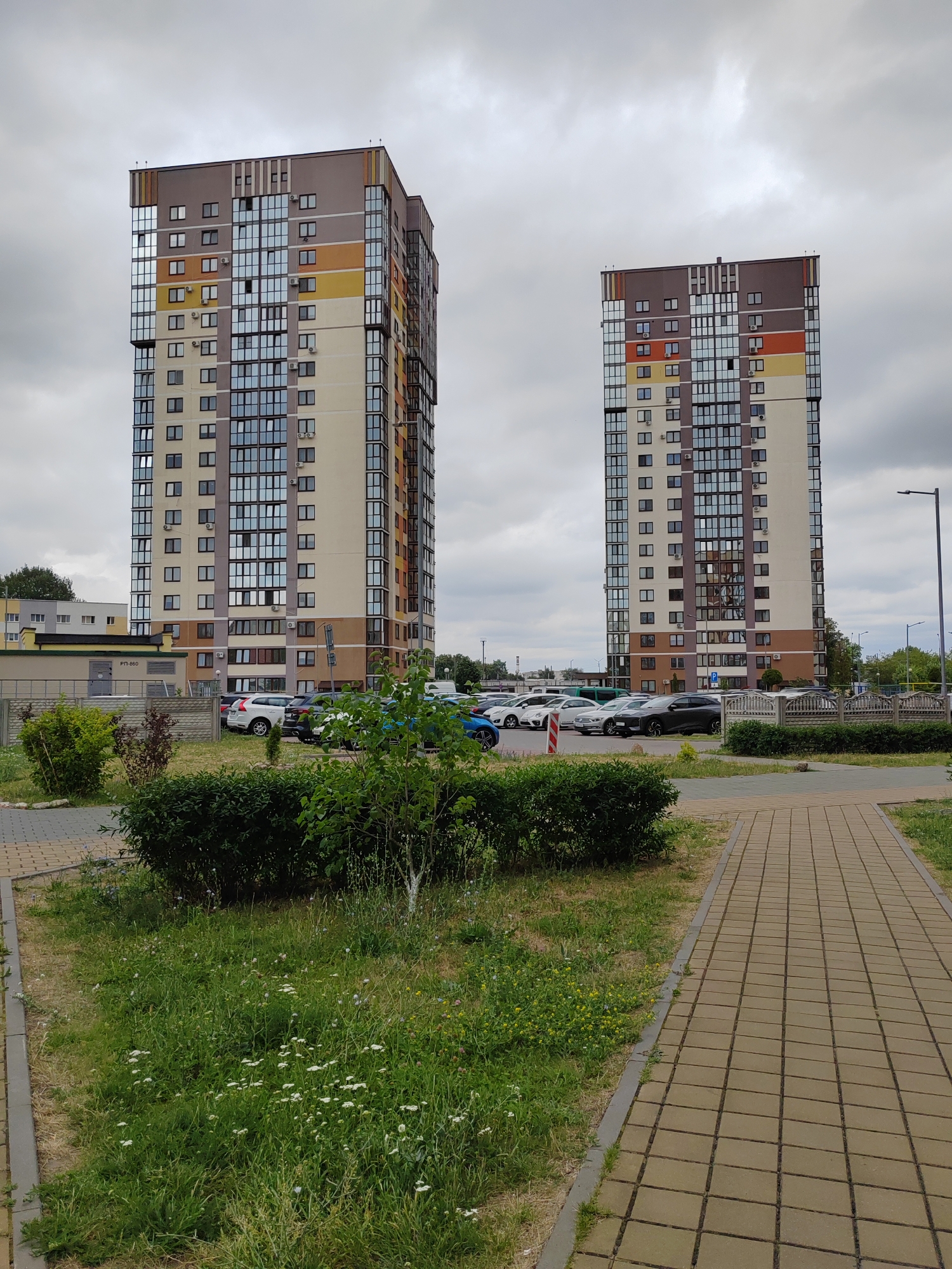 The image shows two modern high-rise apartment buildings in the city of Brest, Belarus, on a cloudy day. The buildings are tall and have a contemporary design with colorful facades featuring horizontal bands of beige, brown, orange, and yellow. Large windows are a prominent feature, giving the buildings a modern and airy appearance. In front of the buildings, there is a parking lot with several cars. A paved walkway runs along the right side of the image, bordered by green grass and bushes. The overall environment suggests a residential area. There are no people visible in the image.