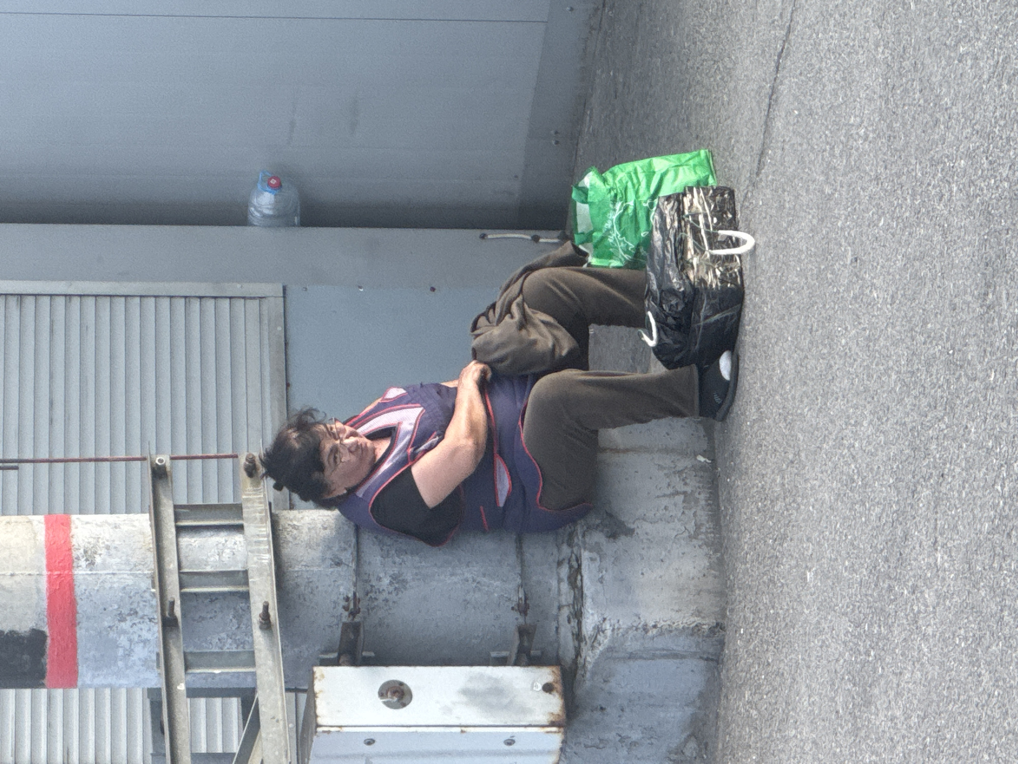 A middle-aged woman wearing a dark shirt, brown pants, a purple vest with dark trim, and black shoes is lying down on a concrete ledge next to a gray building with a metal ladder. A green reusable shopping bag and a black tote bag are positioned near her feet. A large water bottle sits atop the building ledge near the ladder. The scene appears to be outdoors, adjacent to an asphalt surface. The location is Krasnodar, Russia.