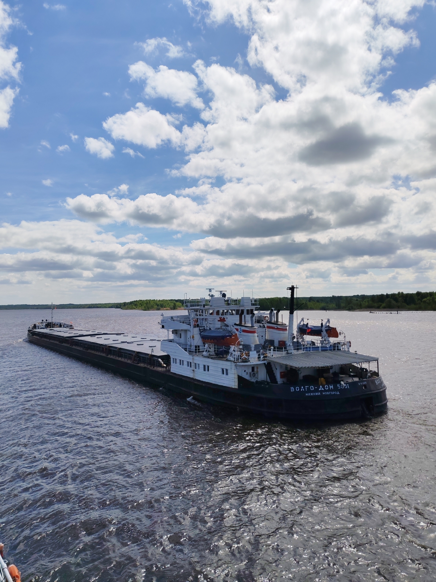 A barge, identified as "VOLGO-DON 5051" from Nizhny Novgorod, is seen on a body of water under a sky with scattered clouds. The barge is dark colored and appears to be a river transport. The background shows a shoreline with green trees. The water is dark and slightly rippled. The image does not appear to be located in Popovo, Russia.