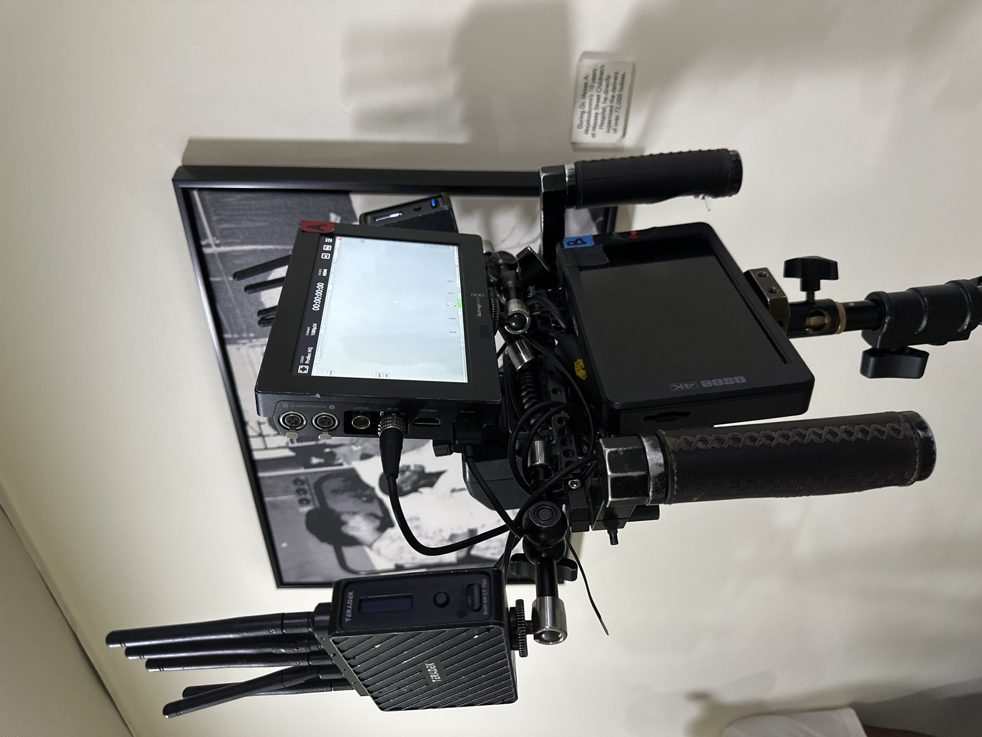 The image shows a video camera rig mounted on what appears to be bicycle handlebars. The rig includes two monitors, one displaying "ProRes HQ" and "1080p24" among other information, and the other displaying "OSEE 4K". A Teradek "Bolt 4K LT 750" transmitter is also attached to the rig.
In the background, there is a framed black and white photograph and a small sign. The sign contains the text "During Dr. Moses A Mojakodunmi's 10 years of Massey Street Children's Hospital, he directly supervised the delivery of over 72,000 babies". Massey Street Children's Hospital is located in Lagos, Nigeria.