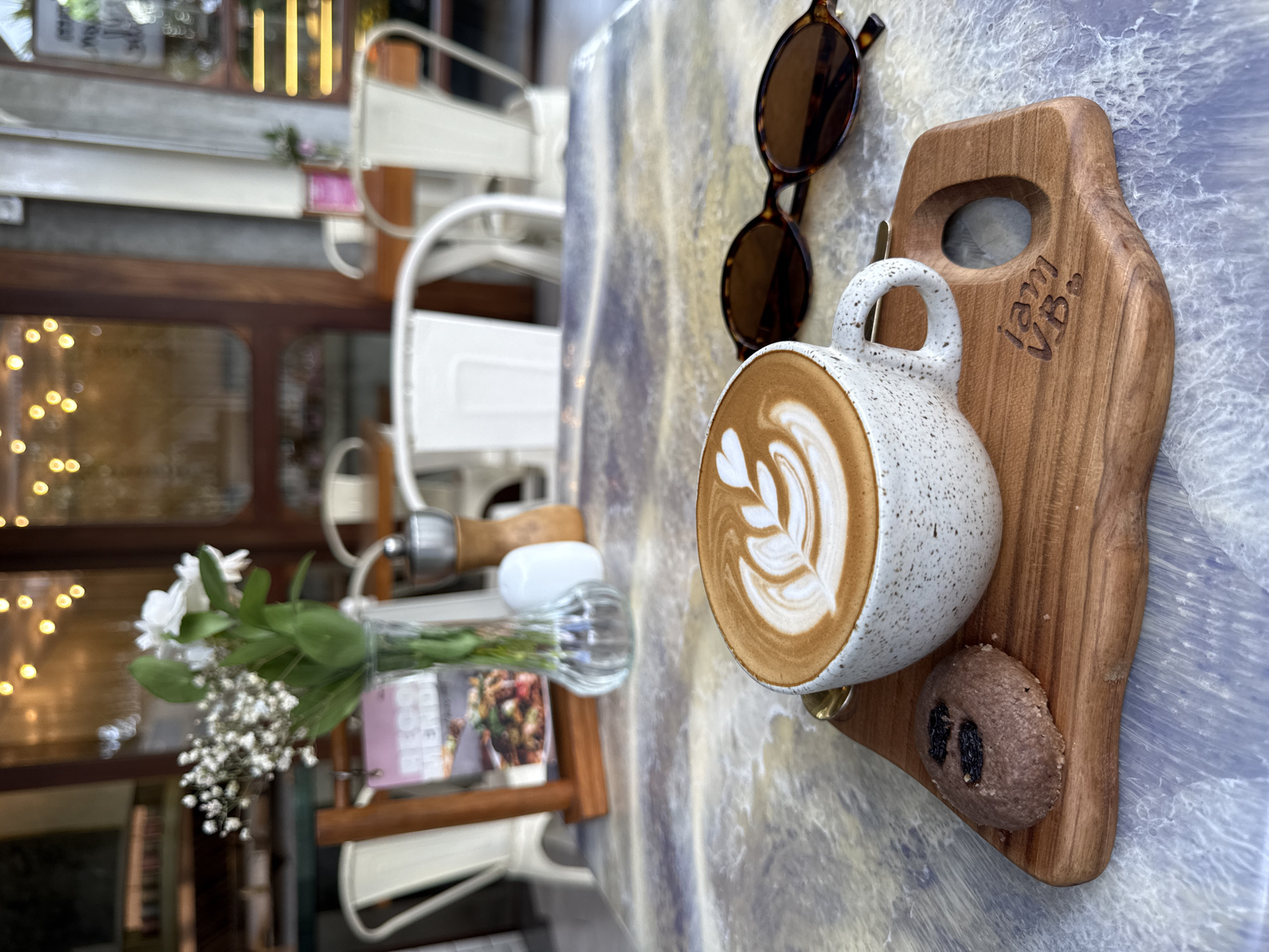 The image shows a latte art coffee on a wooden board next to a pair of sunglasses and a small cookie. The coffee is served in a speckled ceramic mug and has a decorative pattern created in the foam. The wooden board also has an engraving. The scene is set on a table with a light blue surface. In the background, there is a cafe setting with white chairs, a wooden-framed window, and a vase of flowers. It is North Kuta, Indonesia.