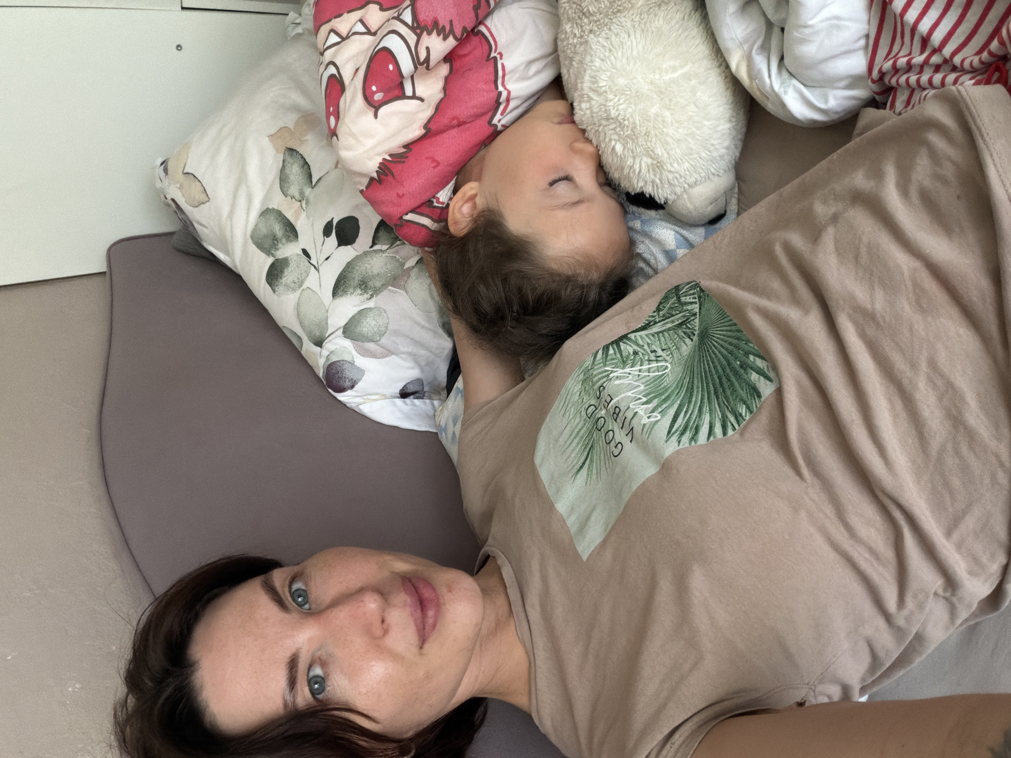 A woman with brown hair and blue eyes lies in bed, facing the camera with a slight smile. She is wearing a tan t-shirt with a graphic print of palm fronds and the words "GOOD VIBES ONLY." Next to her, a young child sleeps, covered by a pink blanket with a cartoon character. The child is resting on a fluffy white stuffed animal and a patterned pillow. The background shows a bed with various pillows and bedding, suggesting a domestic setting. The image appears to be a selfie taken in a bedroom. The location is not specified.
