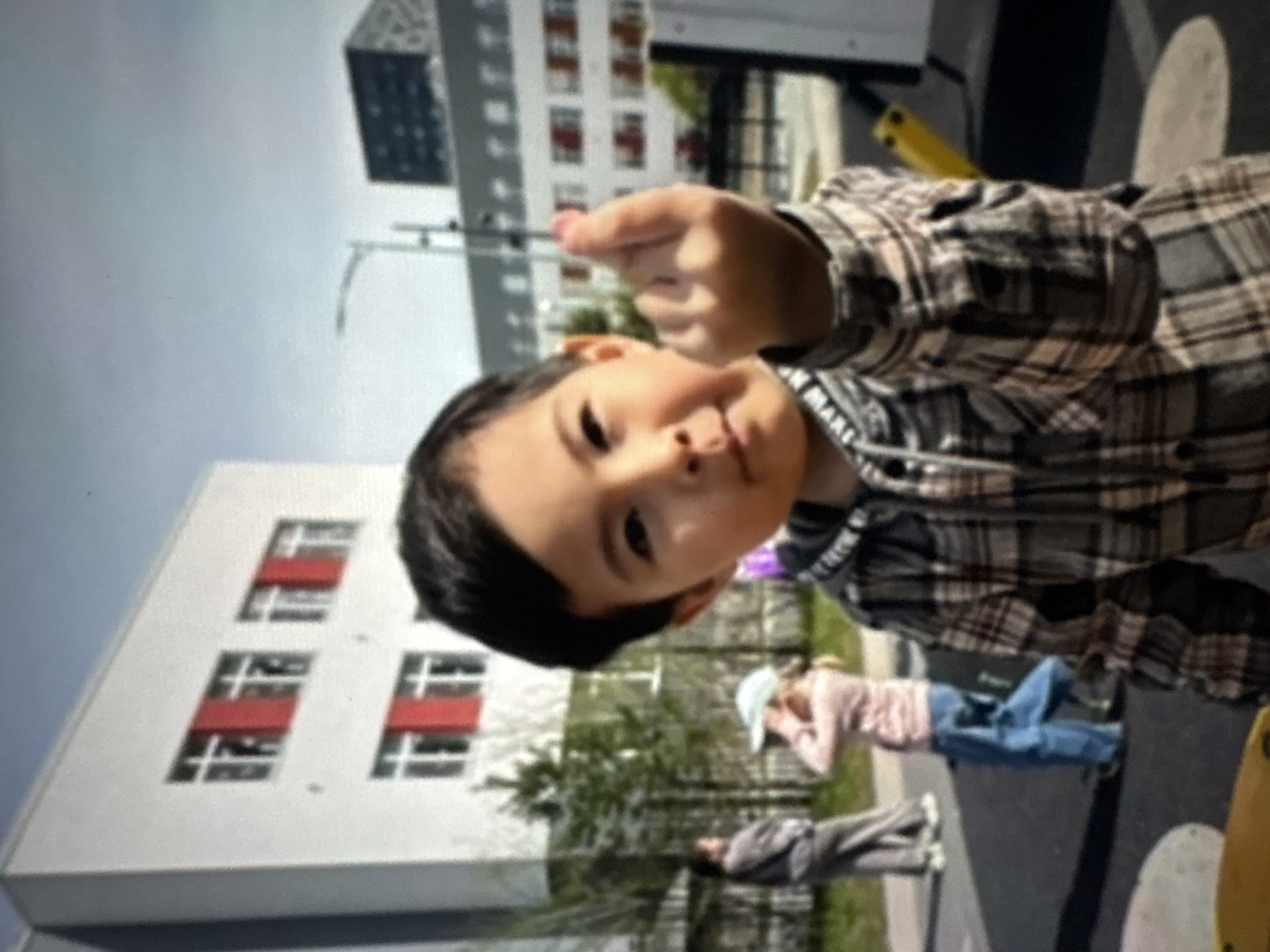 A young child is in the foreground, wearing a plaid shirt and pointing towards the viewer. In the background, there are two buildings with red accents on the windows and a pedestrian walkway with two people. The setting appears to be outdoors, with trees present, suggesting it may be a playground in the city of Ембаево, Russia.