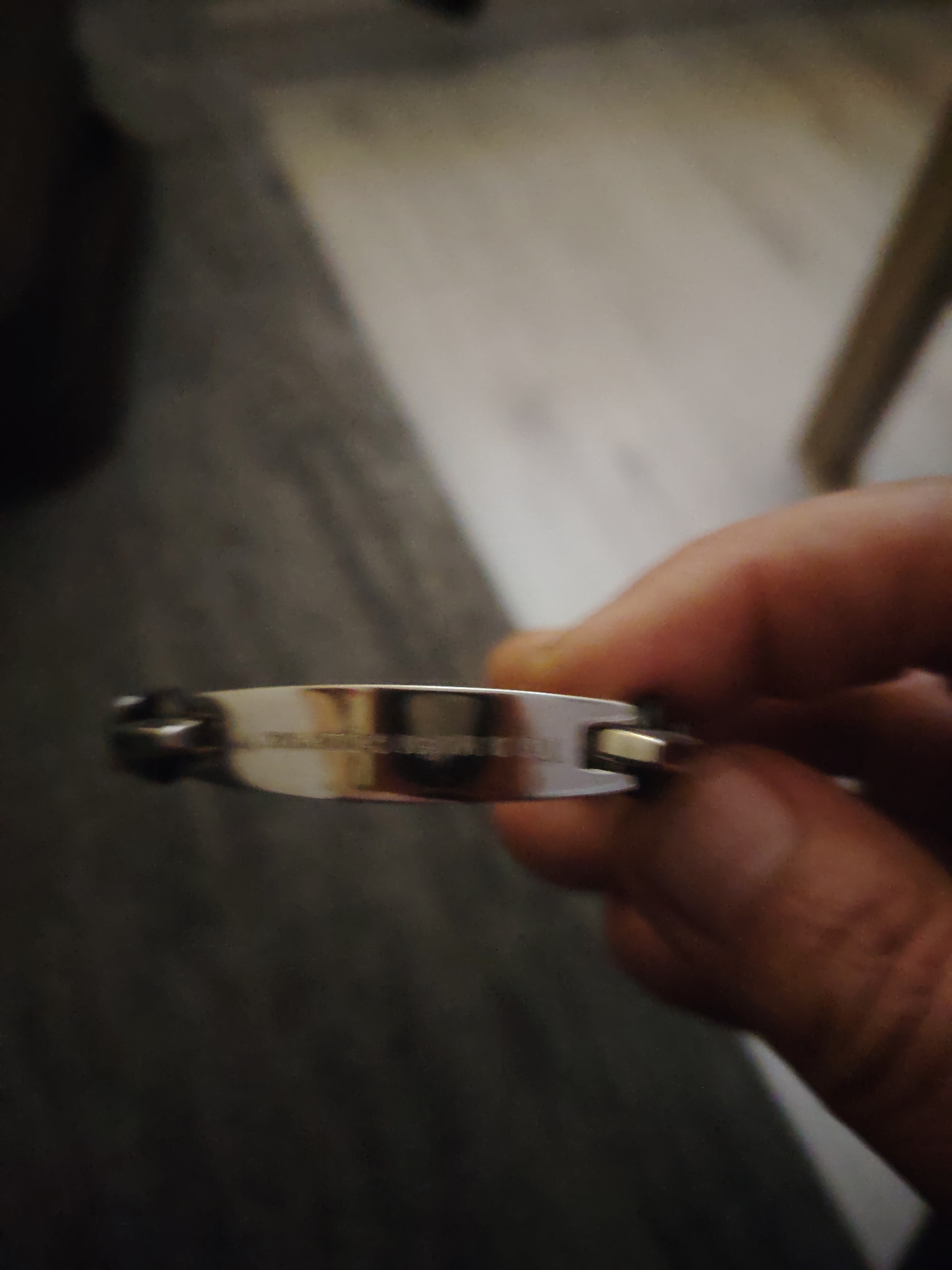A human hand is positioned horizontally, holding a metallic, rectangular bar, consistent with a bracelet, featuring an engraved inscription on its visible surface and a golden-toned clasp detail on its right end. The background is a blurred indoor environment in Huntingdonshire, United Kingdom, characterized by dark, soft textures on the left and lighter, hard surfaces on the right, indicative of common interior spaces.
