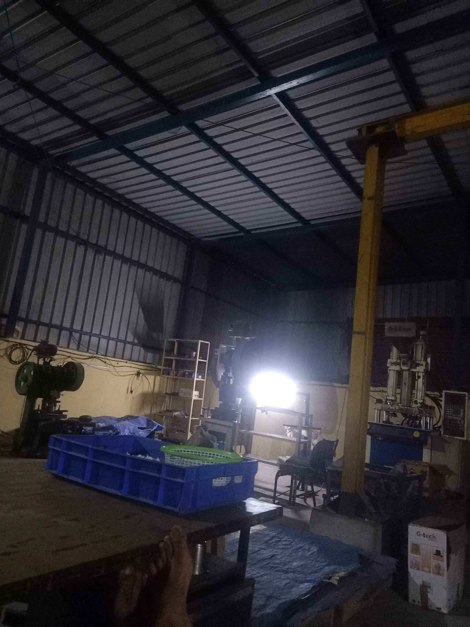 The image shows the interior of a workshop in Dadri, India, likely a manufacturing or assembly facility. The scene is dimly lit, with a corrugated metal roof supported by metal beams, and corrugated metal siding. In the foreground, a person's bare feet are visible resting on a dark, wooden surface, suggesting a moment of rest or inactivity.
To the left, a green industrial machine, possibly a power press, is present. In the center of the image, a large blue plastic crate is filled with some white material and a green mesh basket. Various shelving units and other industrial equipment, including a machine with pneumatic cylinders, are visible in the background. A bright light source emanates from the middle-right section of the image, casting a glow. The floor is partially covered with a blue tarp. A yellow support beam is prominent in the center-right of the frame. The overall impression is of a functional, albeit somewhat cluttered, industrial workspace.
