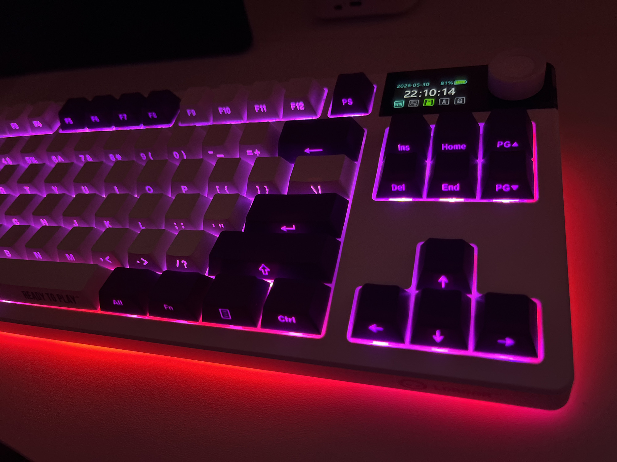 A close-up image shows a white mechanical keyboard with purple and red LED backlighting. The keyboard features a display screen in the upper right corner showing the date "2026-05-30" and time "22:10:14," along with a battery percentage of 81% and other icons. The keyboard has a standard layout, including function keys (F1-F12), alphanumeric keys, modifier keys (Alt, Ctrl), and a numeric keypad. The keys themselves are a mix of white and black, with the black keys being the arrow keys, insert/delete keys, and function keys. The keyboard's bottom edge emits a red light. The location is unknown and there is no information provided that suggests that this image was taken in Varna, Bulgaria.