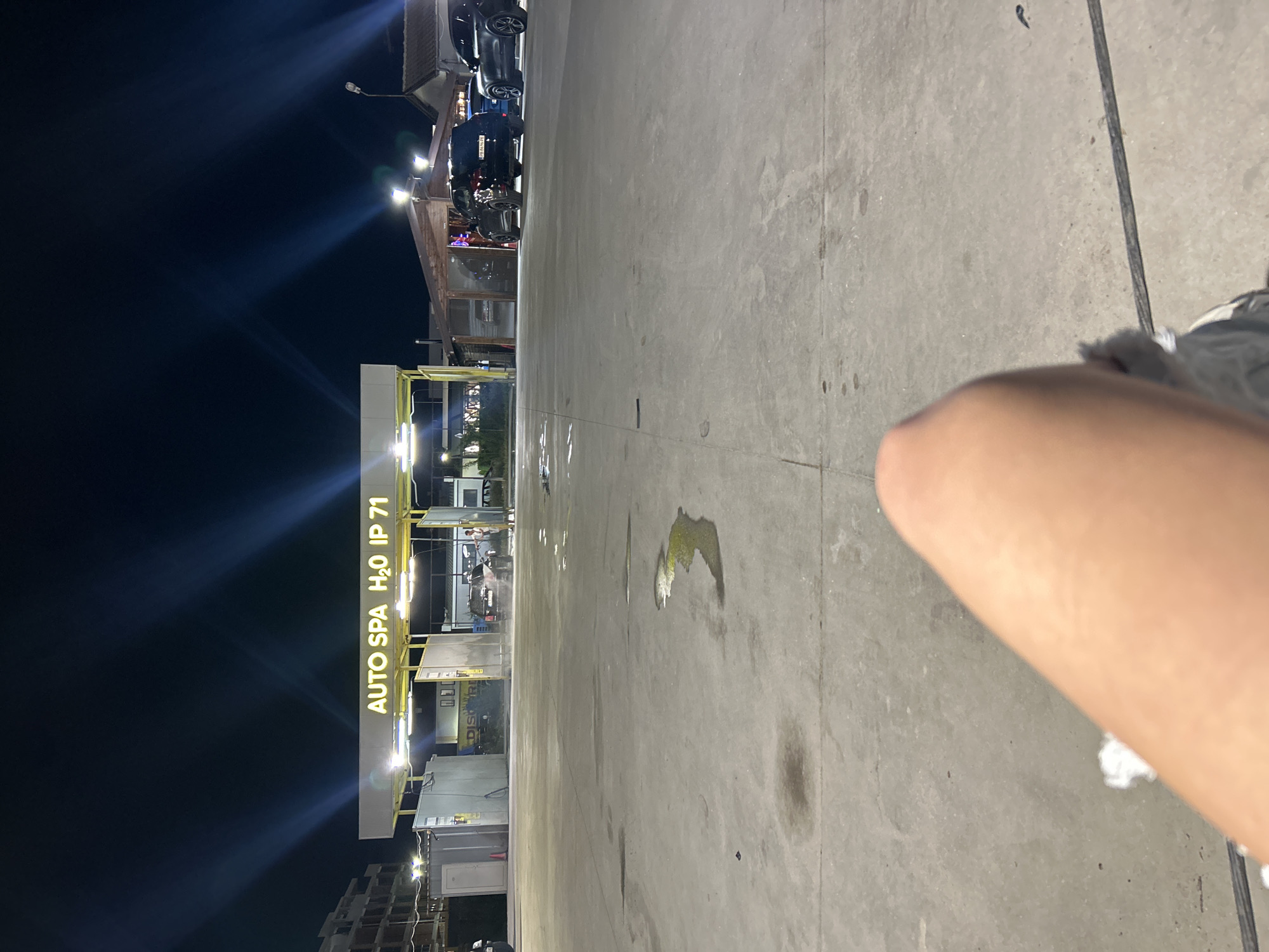 The image depicts an outdoor scene at night in what appears to be a car wash facility. A sign reading "AUTO SPA H2O IP 71" is visible, suggesting a possible business name and location near Plovdiv, Bulgaria. The foreground features a person's arm and possibly a part of their foot. The ground is concrete, marked with stains. Further back, cars are lined up at the car wash area, indicating activity at the facility. The sky is dark and some buildings are visible in the far background.