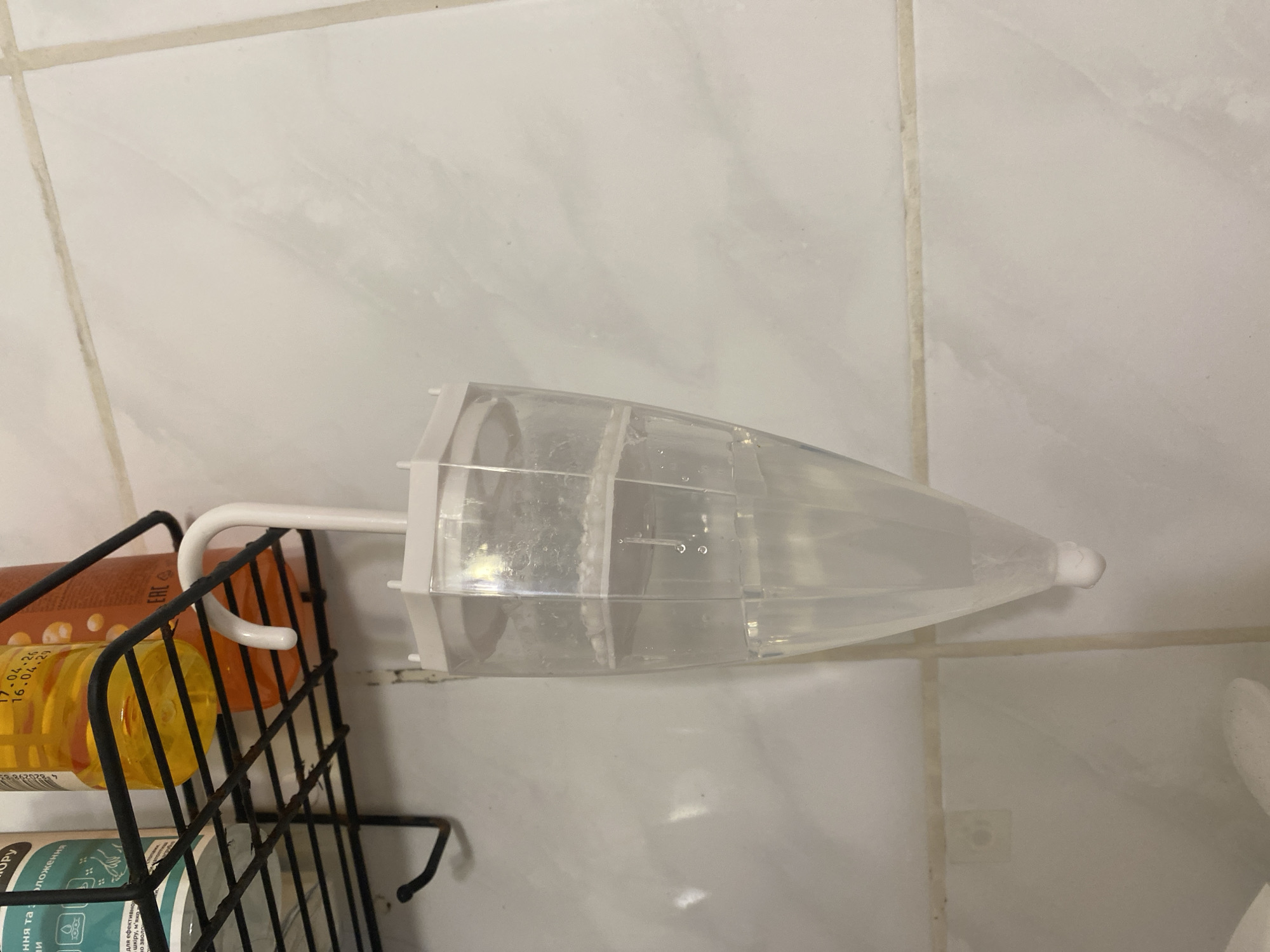 The image shows a close-up of a shower shelf with bottles of toiletries and a clear plastic device shaped like an umbrella hanging from a white hook. The device contains a clear liquid and some small white beads. The background is a tiled wall. The location is the city of Kryvyi Rih, Ukraine. The image does not contain any people or depict any interactions.