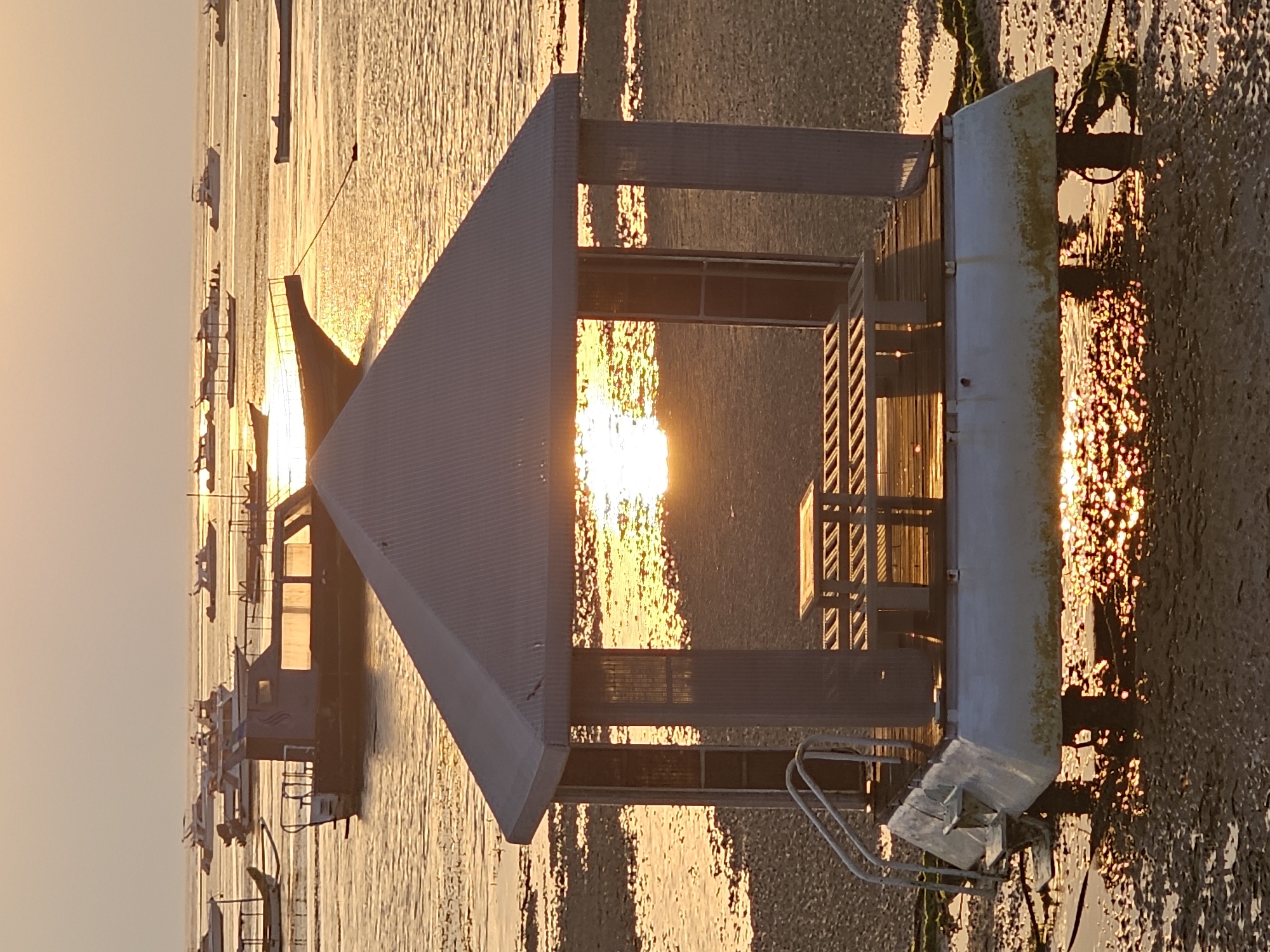 The image captures a serene sunset scene at a beach, likely in or around Denpasar, Indonesia, given the presence of traditional Indonesian fishing boats and a resort-like gazebo. In the foreground, a stilted gazebo stands on a floating platform, partially submerged in shallow water. The gazebo has a simple, weathered design with a corrugated metal roof and vertical slatted walls, framing a wooden deck with two benches. Metal railings and a ladder lead up to the platform. The setting sun, a brilliant orb of light, is visible through the gazebo's opening, casting a warm, golden glow on the water and the sandy, muddy shore.
In the background, numerous boats are anchored in the sea. These range from smaller, traditional wooden fishing boats with outriggers to larger, modern motorboats and yachts. The water is calm, reflecting the warm hues of the sunset sky. The overall atmosphere is tranquil, indicative of a peaceful evening by the sea. There are no people visible in the image.
