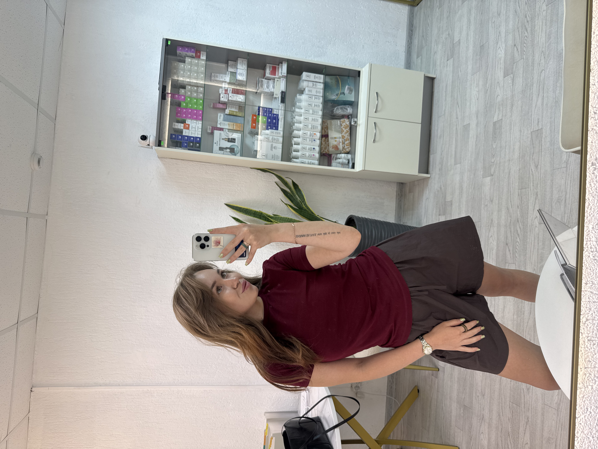 A young woman with long, light brown hair, wearing a burgundy t-shirt and dark brown shorts, takes a selfie in a well-lit room. She is looking at her phone, which she holds in her right hand. Her left hand rests on her shorts. She is wearing a watch on her left wrist and a ring on her left index finger. Her nails are painted a light green color.
In the background, to the right of the woman, is a tall, white display cabinet filled with various cosmetic products. The cabinet has glass doors and shelves, and is illuminated from above. Above the cabinet, a small white security camera is mounted on the wall. To the left of the woman, a round table with a white top and gold legs is visible, with a black bag placed on it. Behind the table, a white wall with a textured surface is seen. A potted plant with long, green leaves stands next to the display cabinet. The floor is covered with light-colored wooden planks. The scene appears to be an interior space, possibly a beauty salon or a retail store. The lighting is bright and even. There is no indication of the specific location within Chaikovsky, Russia.
