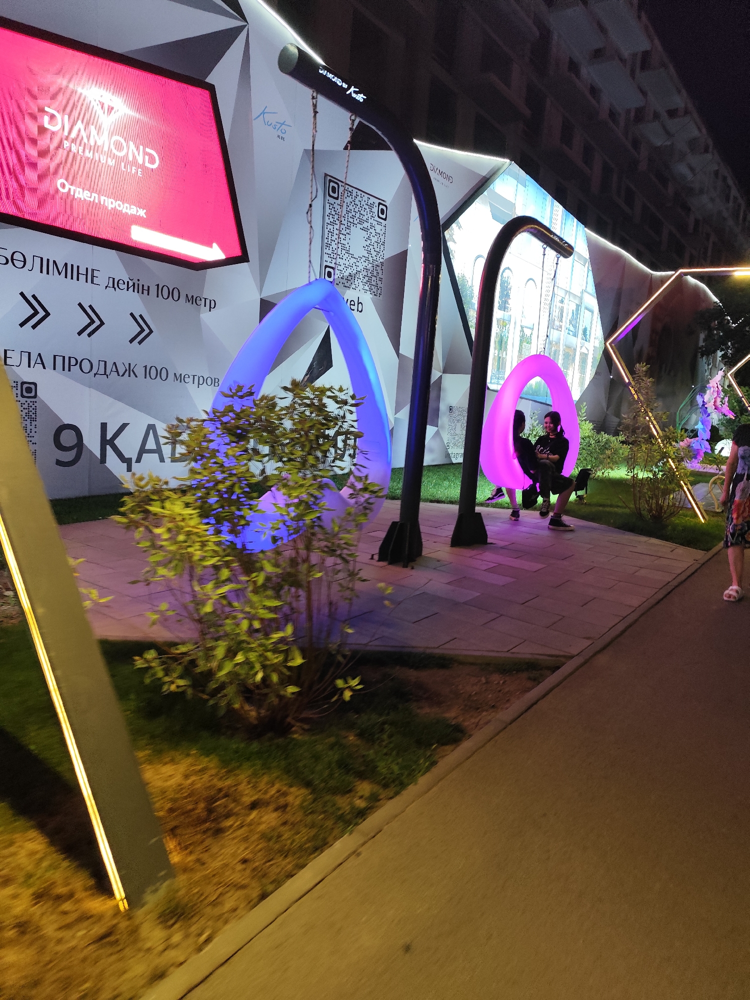 An outdoor nighttime scene in Almaty, Kazakhstan, features a modern, geometric light installation. The installation includes large, illuminated rings in blue and pink, one of which is occupied by two individuals, a woman and a man, seated facing each other and appearing to be conversing. Behind the installation, a building with multiple stories and windows is visible, suggesting an urban setting. Promotional signage with text in what appears to be Cyrillic script is displayed prominently. The ground is paved with interlocking stones and is illuminated by spotlights and the glowing rings. A person in a floral dress is walking away from the camera on the right side of the frame. The overall atmosphere is vibrant and contemporary.