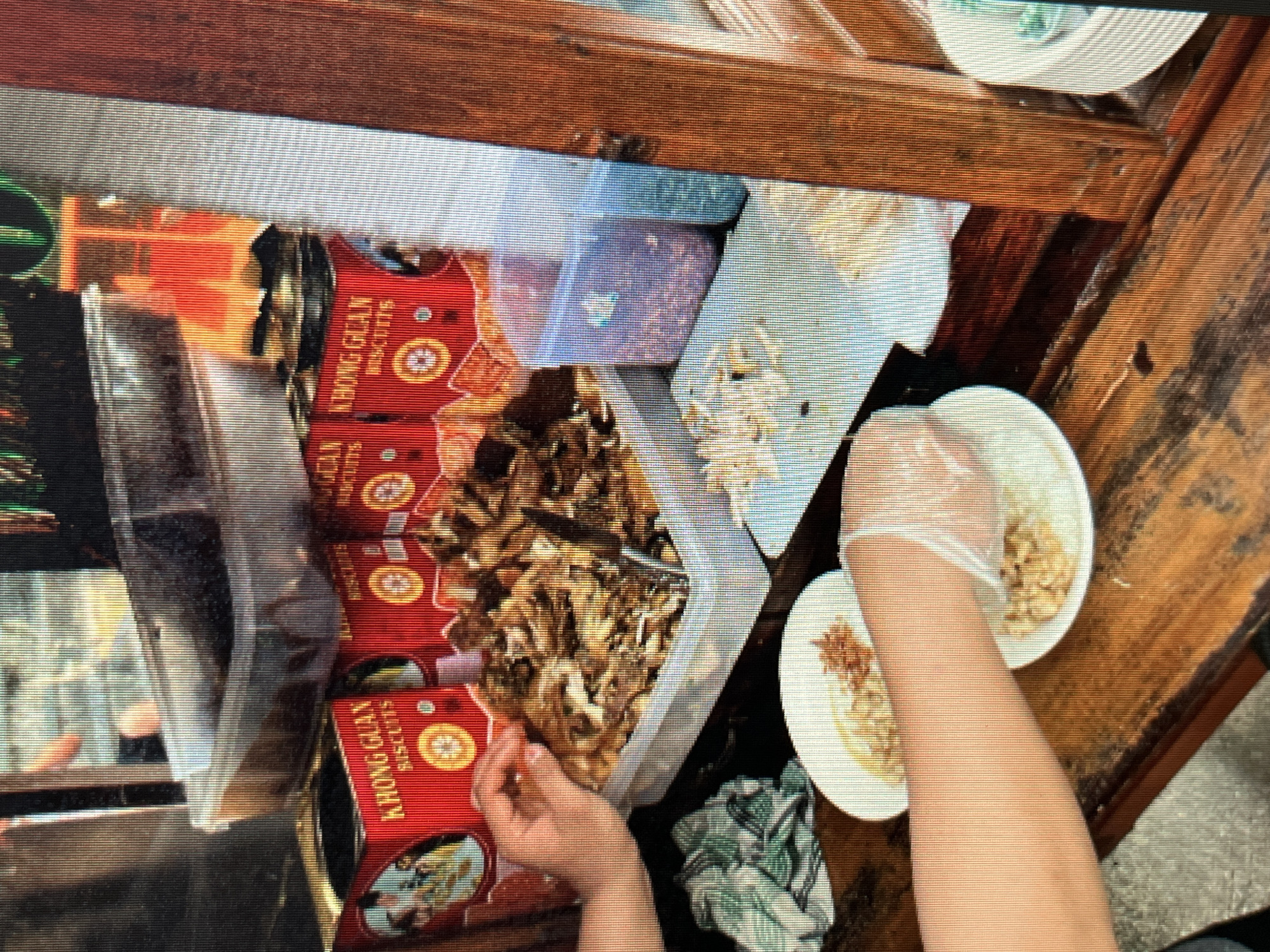 This image captures a scene at a street food stall in Sejahtera, Indonesia. The stall features a wooden counter and a display of various food items and containers. In the foreground, two white plates are visible, one with food that appears to be rice or noodles topped with fried shallots, and another with a portion of shredded chicken. A person's arm, wearing a transparent glove, is seen reaching towards the second plate, suggesting they are preparing or serving food.
Behind the plates, a large white plastic container holds a generous portion of what appears to be crispy fried snacks or noodles. To the left of this container are several red metal cans labeled "KHONG GUAN BISCUITS" in prominent white lettering. These cans are stacked, with one in the front showing a circular emblem with a drawing of people.
Further back, a clear plastic container filled with a reddish-brown granular substance is placed next to another translucent blue container. The background is somewhat blurred, but hints of other food items or ingredients in larger containers are discernible. The overall environment suggests a busy outdoor food vending operation. The lighting indicates it could be daytime, with artificial light possibly contributing to the illumination. The texture of the wooden counter suggests it is well-used.