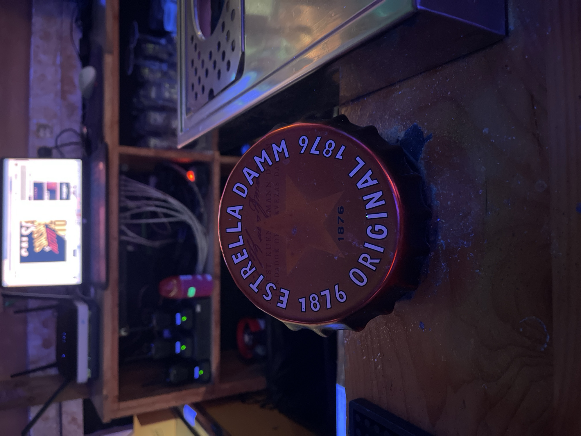 The image depicts a close-up view of a bottle cap, likely from a beer, placed on a wooden surface, possibly a bar top. The cap is orange with white lettering that reads "ESTRELLA DAMM ORIGINAL 1876". In the background, a laptop is visible on a wooden shelf, along with networking equipment and a pink object that could be a bottle of liqueur. The lighting is dim, with some blue and purple hues. The scene appears to be indoors, in a setting that suggests a bar or a similar establishment in Barcelona, Spain, given the brand of beer.