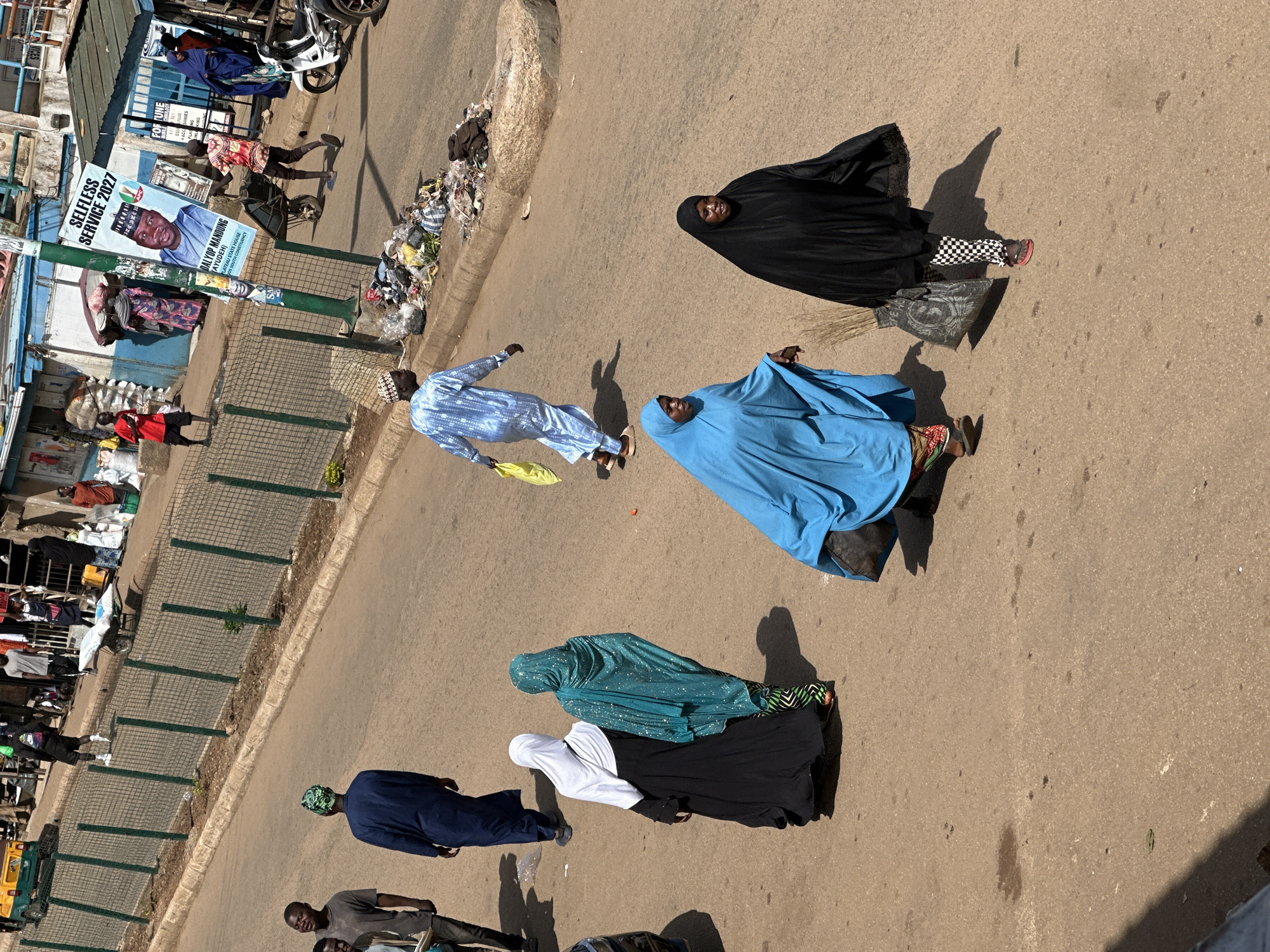 The image depicts a street scene in Bukuru, Nigeria. In the foreground, three women are walking across a paved road. The woman on the left wears a bright blue abaya and a patterned skirt, carrying a dark bag. The middle woman is partially visible, wearing a black abaya and patterned leggings, and carrying a broom. The woman on the right wears a black abaya and patterned leggings, also carrying a broom. Behind them, two other women are walking away from the camera, one in a white hijab and black abaya, the other in a turquoise hijab and patterned skirt. Further in the background, several men are walking, including one in a blue robe and cap. A poster featuring a man's portrait with the text "SELFLESS SERVICE 2027" is attached to a lamppost. Various objects are scattered along the roadside, including trash and a wheelbarrow. A white motorcycle is parked on the right side of the street. The environment appears to be a busy urban area with buildings and a wire fence lining the street. The lighting suggests it is daytime with bright sunlight, casting shadows on the ground.