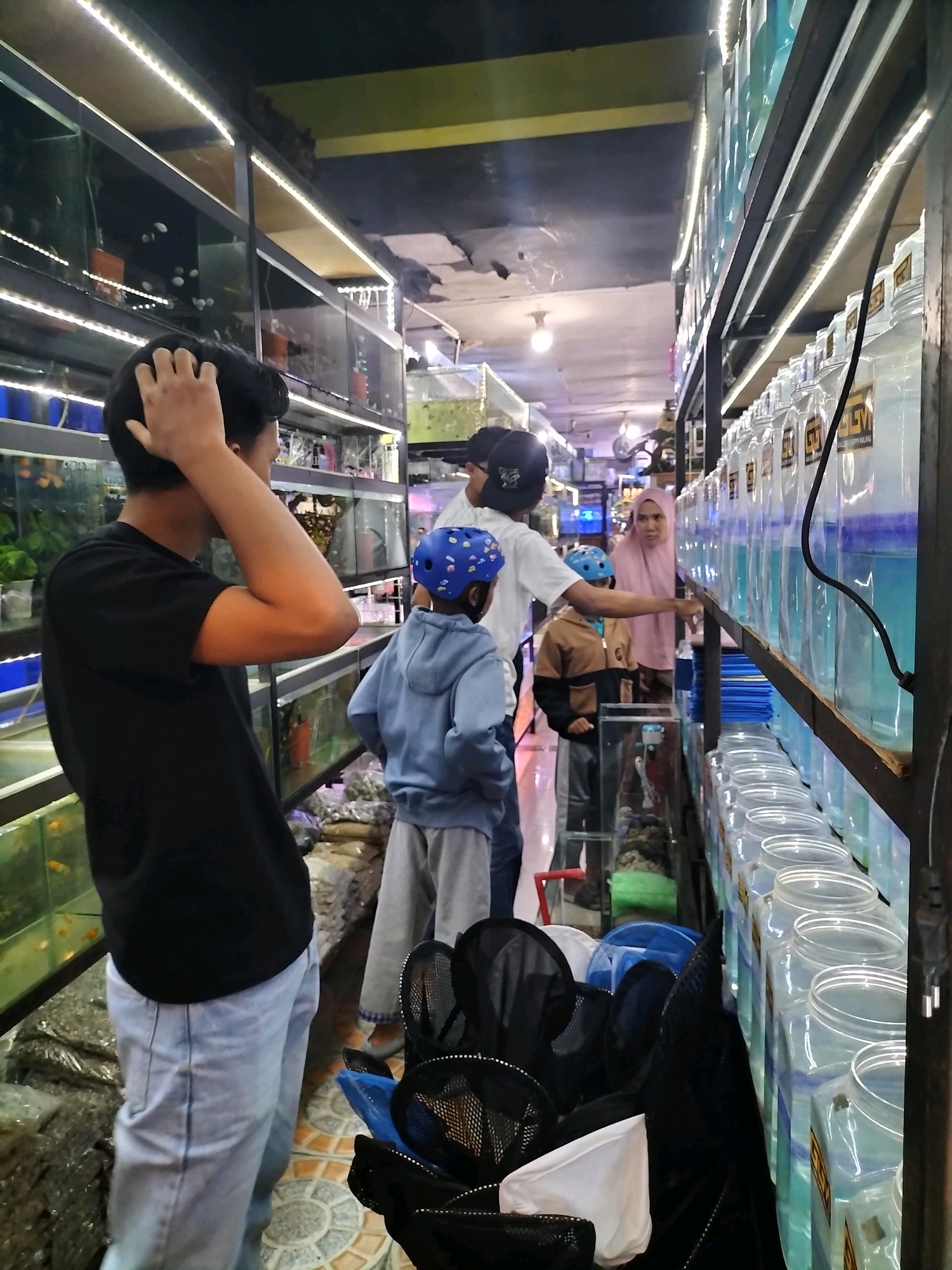 In the city of Malang, Indonesia, a busy pet store selling fish is captured in this image. Rows of clear plastic containers filled with blue liquid and fish are stacked on shelves, illuminated by LED strip lights. A person in a black t-shirt and light blue jeans stands in the foreground, facing away from the camera and scratching their head. To their right, a child wearing a blue helmet and a light blue hoodie stands, looking towards the fish containers. Another child in a brown jacket and grey pants is also present, along with a woman wearing a pink hijab. Bags of gravel and aquarium supplies are visible on the floor. The overall impression is a bustling market scene focused on aquatic pets.