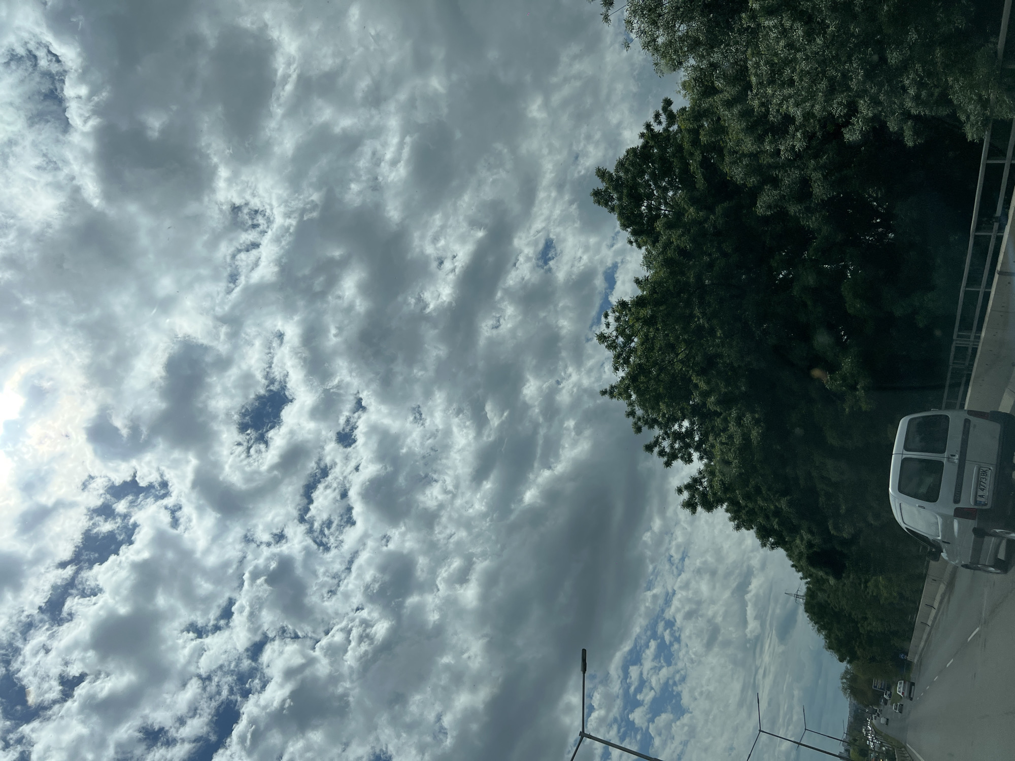The image shows a cloudy sky above a road in Burgas, Bulgaria. The sky is filled with white and gray clouds with patches of blue. Green trees line the right side of the road. A white van is on the road. Streetlights are visible on the left. Other vehicles are in the distance.