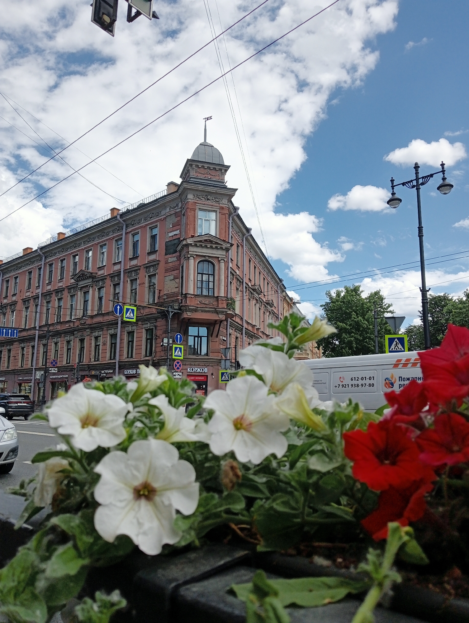 The image captures a street scene in Saint Petersburg, Russia, with a focus on an ornate, multi-story building with a distinctive corner tower. The building, painted in a faded pink hue, features numerous windows and decorative architectural elements. In the foreground, a planter box overflows with white and red petunias, partially obscuring the street and vehicles. Across the street, a white van is visible with Russian text and contact information printed on its side. A lamppost stands to the right, and overhead, traffic signals and power lines crisscross the sky, which is dotted with white clouds against a blue backdrop. Road signs and crosswalk markings are also present on the street.