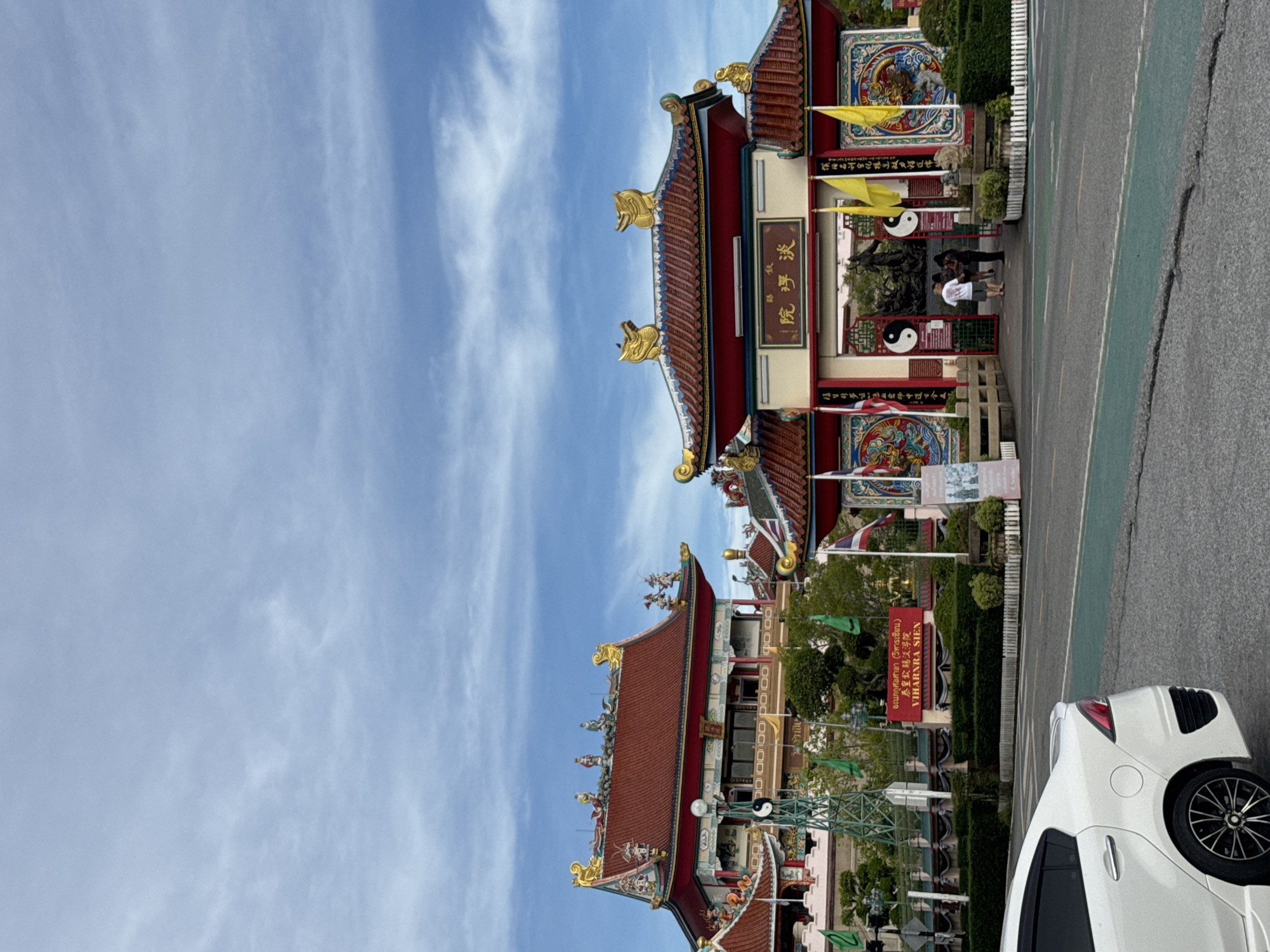 Here is a description of the image:
The photograph, taken in daylight, captures a vibrant scene in มาบฟักทอง, Thailand, focusing on a Chinese temple known as Viharnra Sien, also referred to as "Anek Kusala Sala (Viharn Sien)." The temple features traditional architecture with red roofs adorned with golden ornaments. The entrance displays intricate decorative details, including Yin-Yang symbols and signage in both Thai and Chinese characters, with the Thai text translating to "Anek Kusala Sala (Viharn Sien)" and "Viharnra Sien."
The foreground includes a portion of a white car, parked on the right side of the frame, which appears to be in the parking area in front of the temple. The temple stands alongside a road or parking lot. The sky is predominantly blue with scattered clouds, indicating fair weather. Several people can be seen near the temple entrance.