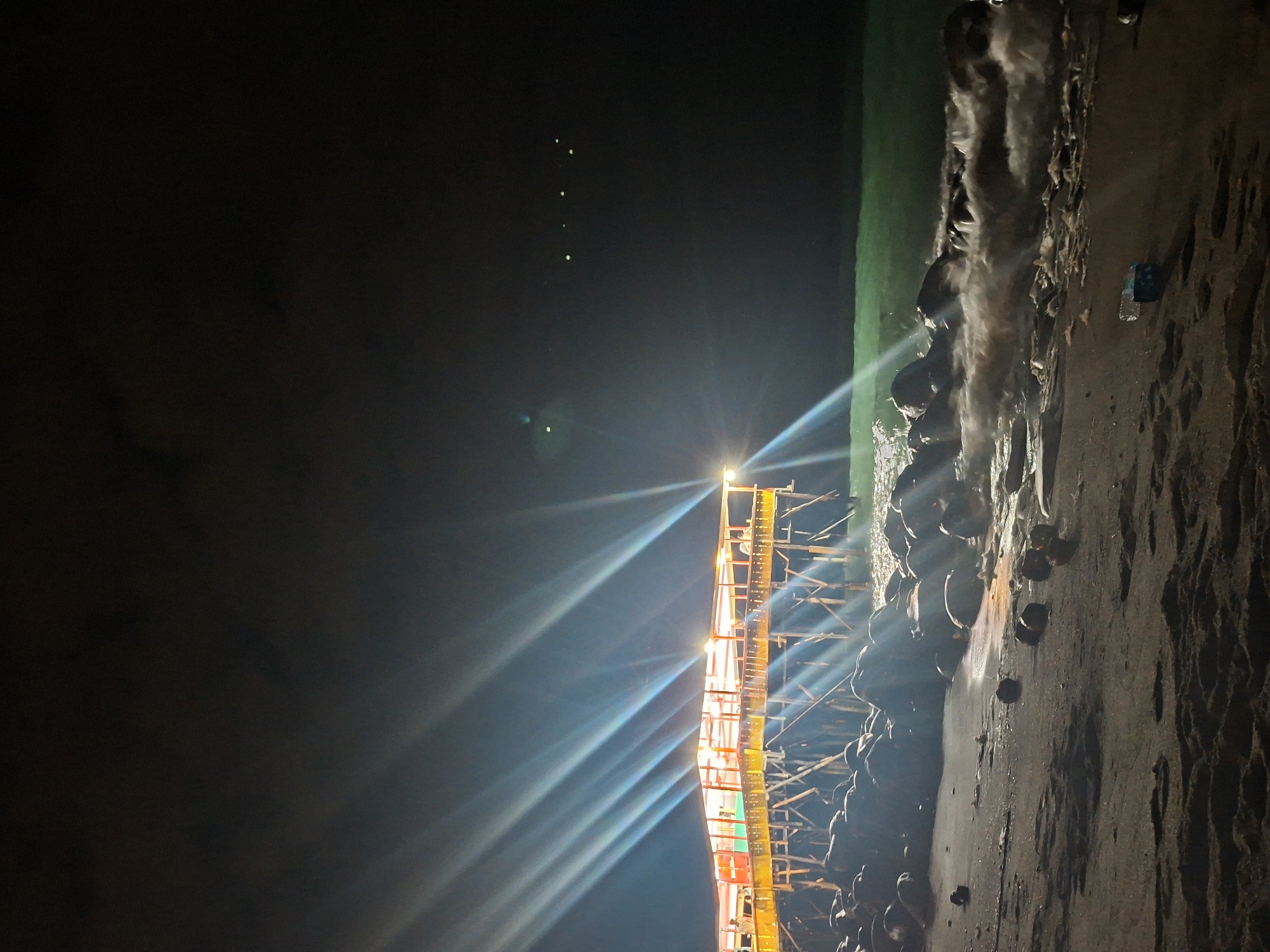 The image shows a pier in Lhokseumawe, Indonesia, at night. The pier is brightly lit, and the light reflects off the water. Waves crash against the rocks and the shore. The beach is dark and wet. A single water bottle is visible on the sand. The sky is dark with a few dim lights visible.