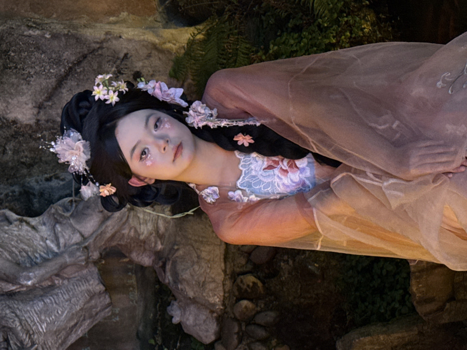 An individual, appearing female and of East Asian descent, is depicted seated in an outdoor cultural or garden setting within Yuzhong District, China. The subject is centrally positioned, facing slightly right with a direct, neutral gaze towards the viewer. She wears a traditional Chinese-style ensemble, including a light blue top with intricate pink and white floral embroidery, overlaid with a translucent, light peach-colored sheer garment that drapes over her arms and torso. Her dark hair is styled in an elaborate updo, adorned with multiple artificial pink and white flowers and delicate beaded accessories. A long, dark braided section of hair extends over her right shoulder, also decorated with small pink flowers. Her facial makeup includes pink eyeshadow and small, star-shaped glitter applied beneath the outer corners of her eyes. The background features a large, rough-hewn stone formation or sculpted rock face to the upper left, with a discernible small stone animal figurine visible at its base. Dense green foliage is present in the lower right and background. The scene is illuminated by ambient light, characteristic of dusk or evening conditions, with focused lighting on the subject.