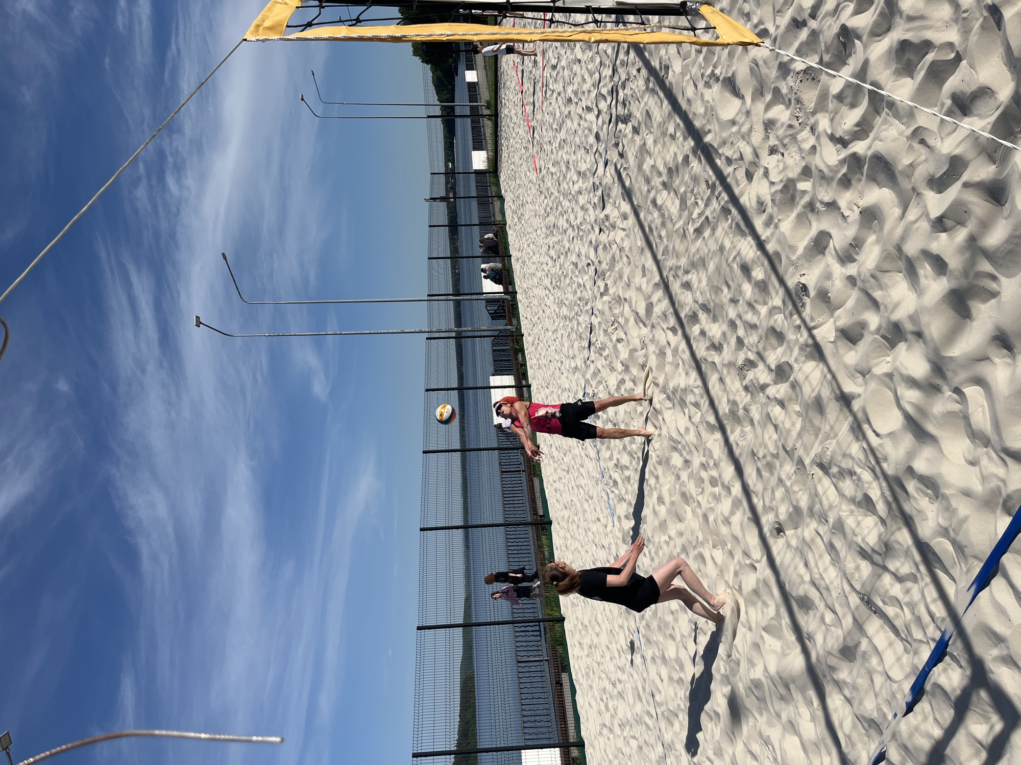 The image depicts a beach volleyball game in progress on a sandy court. Two individuals are visible on one side of the net. One is in mid-air, striking a volleyball. The other player is on the ground, seemingly in a defensive position. The backdrop includes a fence, water, and light poles. The image was taken in the city of Chaikovsky, Russia.