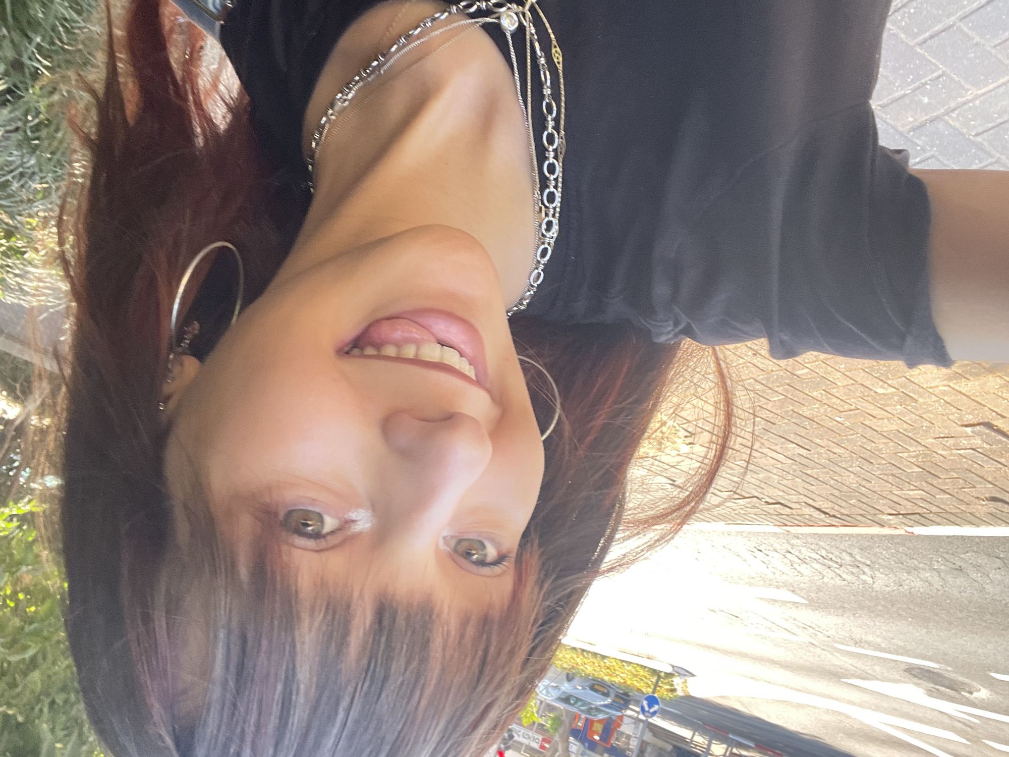 A woman with dark brown hair and bangs is taking a selfie outdoors. She is wearing a black t-shirt and several silver necklaces. She has on large hoop earrings. The woman is smiling with her mouth open, showing her teeth. Her head is tilted to the left.
In the background, sunlight is illuminating a brick sidewalk and a street. There are some trees or foliage visible at the top of the frame. A blue traffic sign with a white arrow is visible on the left side of the image, along with a car. This scene is located on a street in Raanana, Israel, likely during daylight hours.