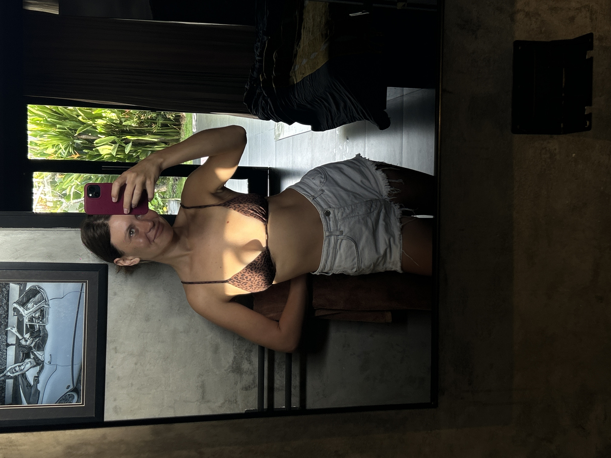 A woman is taking a selfie in a mirror. She is wearing a leopard-print bikini top and white denim shorts. The location appears to be Mengwi, Indonesia, with tropical foliage visible through a window. A framed black and white picture of a car hangs on the wall. A black fixture is visible on the wall behind the woman. A towel is draped over what looks like a towel rack. A black curtain hangs above the woman.